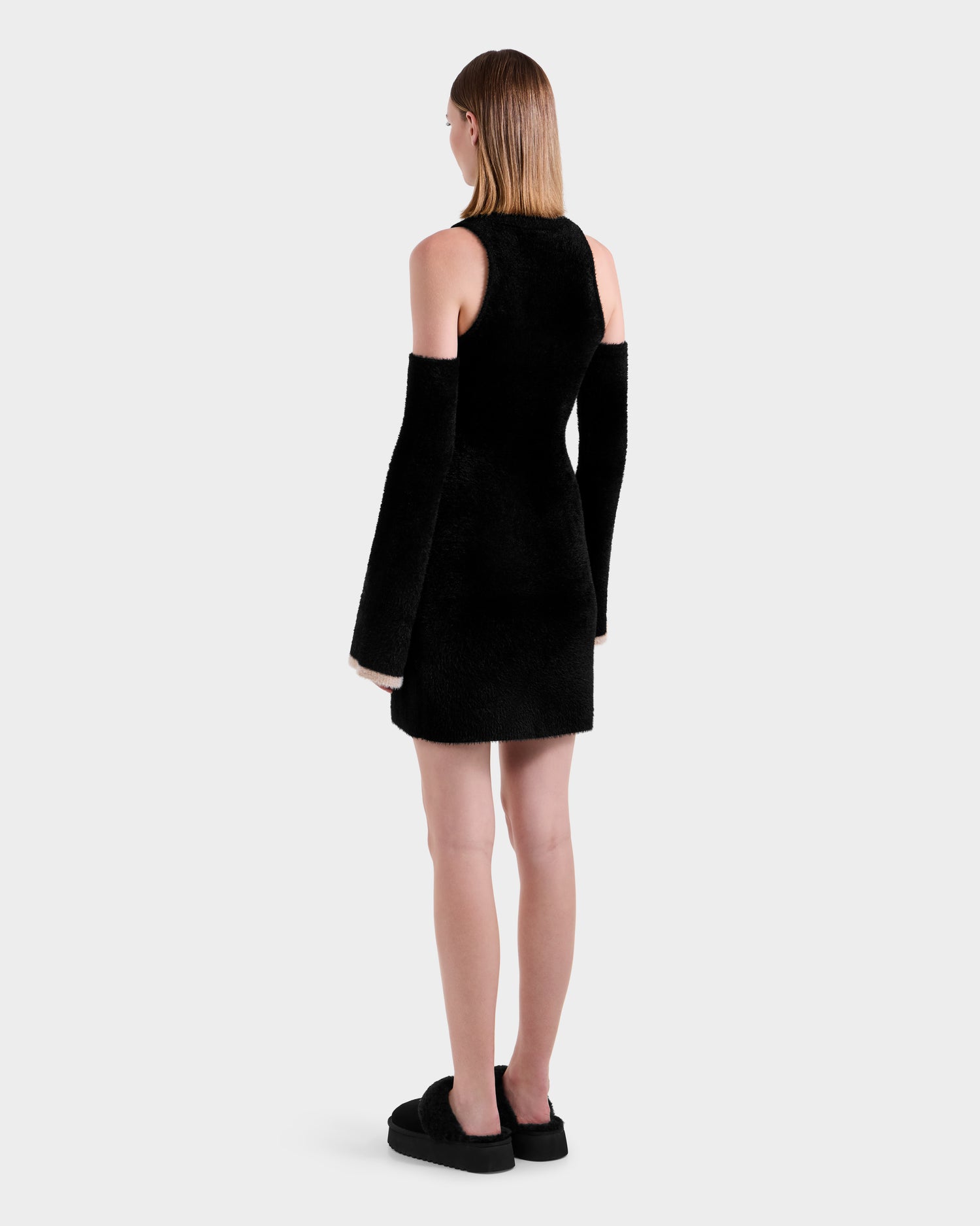A woman with straight hair tucked behind her ears wears the Rudsak ZARAH - 8525696 Black fuzzy knit dress with sleeveless black fur, matching long detached sleeves, and black platform shoes. The background is plain light gray.