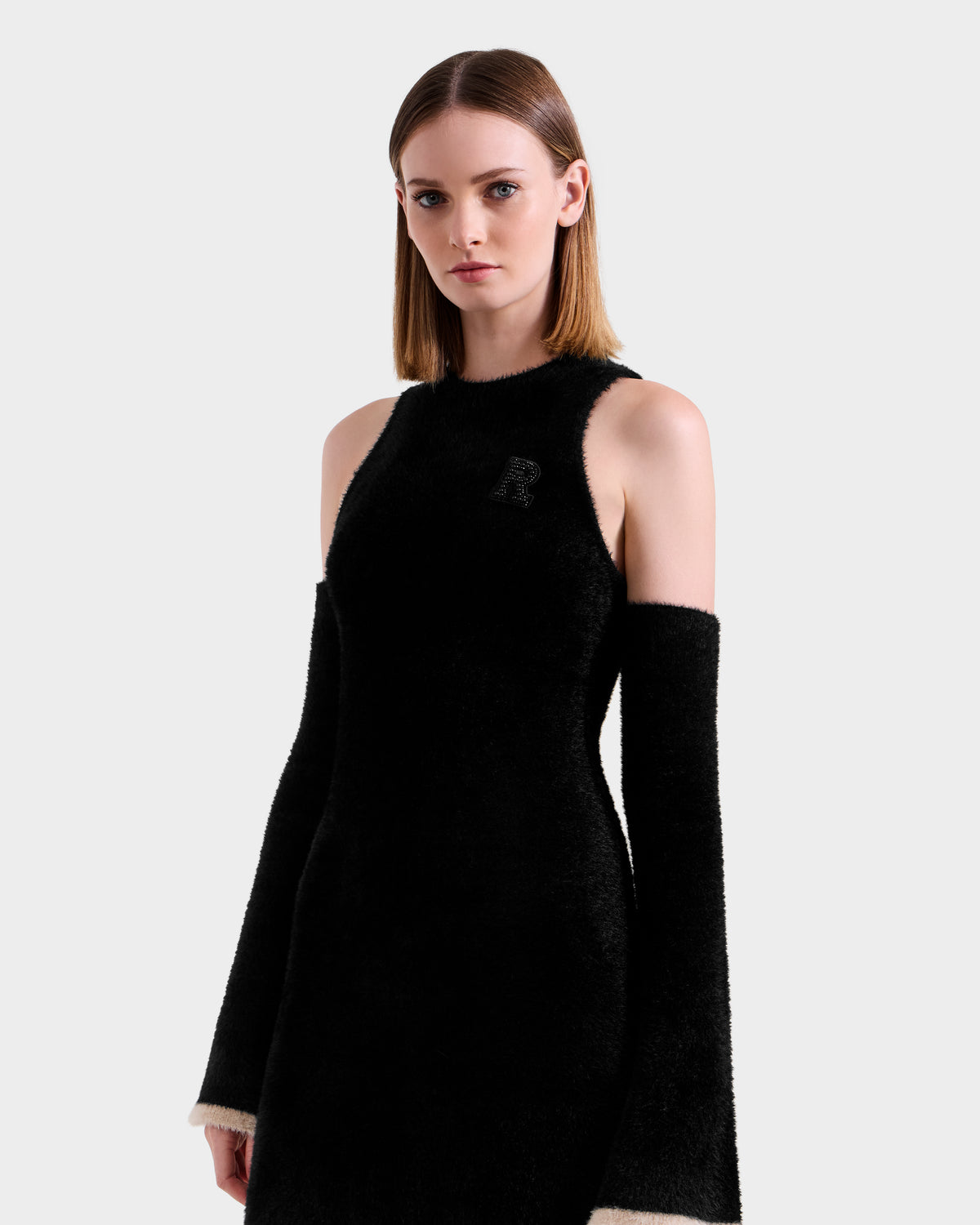 A woman with straight, shoulder-length hair models the Rudsak ZARAH - 8525696 Black—a versatile sleeveless dress with detachable long sleeves and white cuffs—standing against a plain light grey background.
