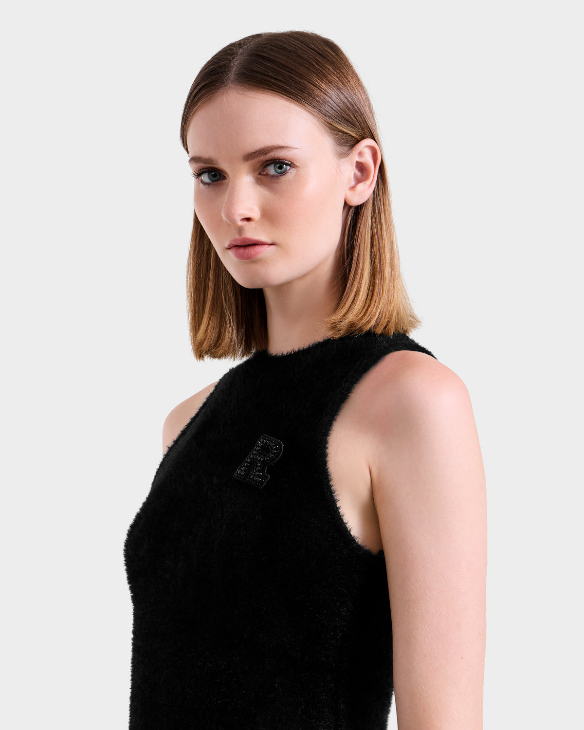 A woman with straight, shoulder-length light brown hair models the Rudsak ZARAH - 8525696 Black sleeveless textured top featuring a decorative R on the chest, posing against a plain light gray background.