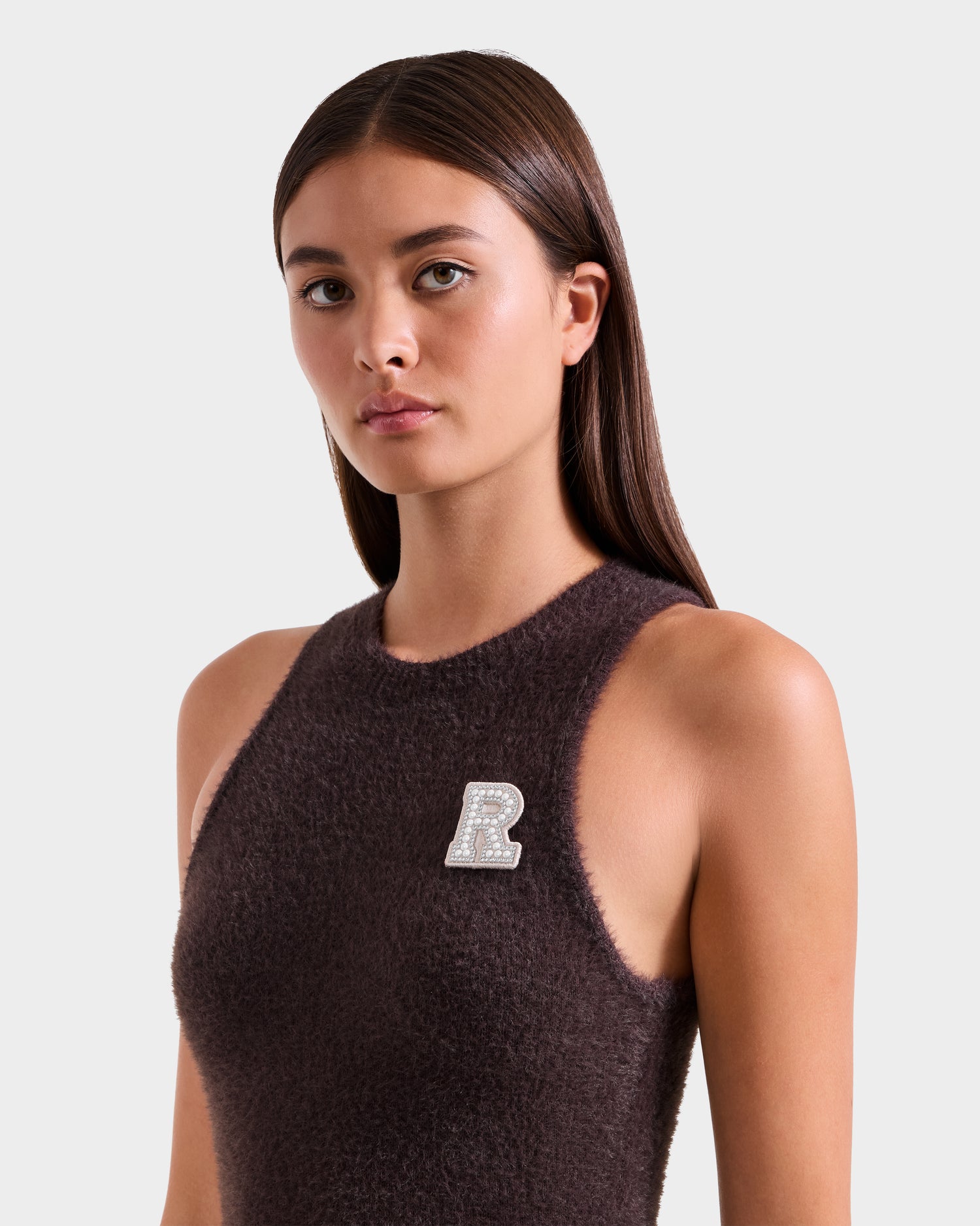 A woman with straight brown hair models the ZARAH - 8525696 Espresso sleeveless fuzzy knit dress by Rudsak, featuring a sparkly "R" brooch on her chest, against a plain light background.