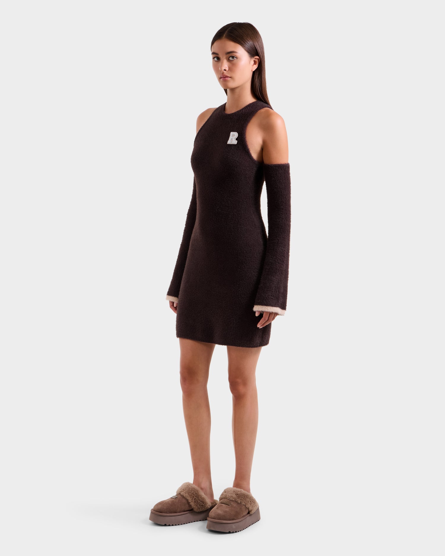 A woman models the Rudsak ZARAH - 8525696 Espresso, an après-ski-inspired dark brown one-shoulder knit dress with a white B patch and beige fuzzy slippers, standing against a plain white backdrop.