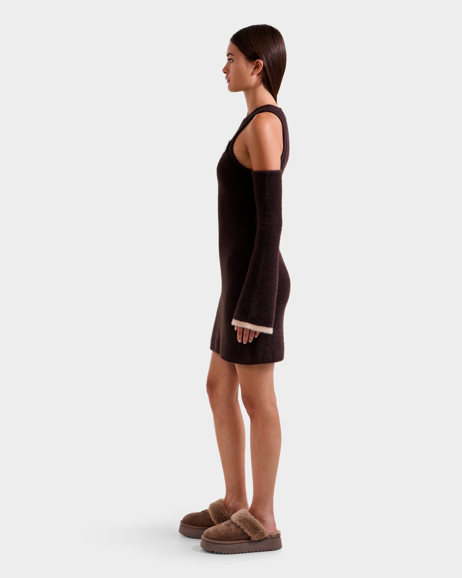 A woman stands in profile against a plain background, wearing the Rudsak ZARAH - 8525696 Espresso sleeveless fitted dress with detached sleeves and tan fuzzy slippers, channeling cozy après-ski style.