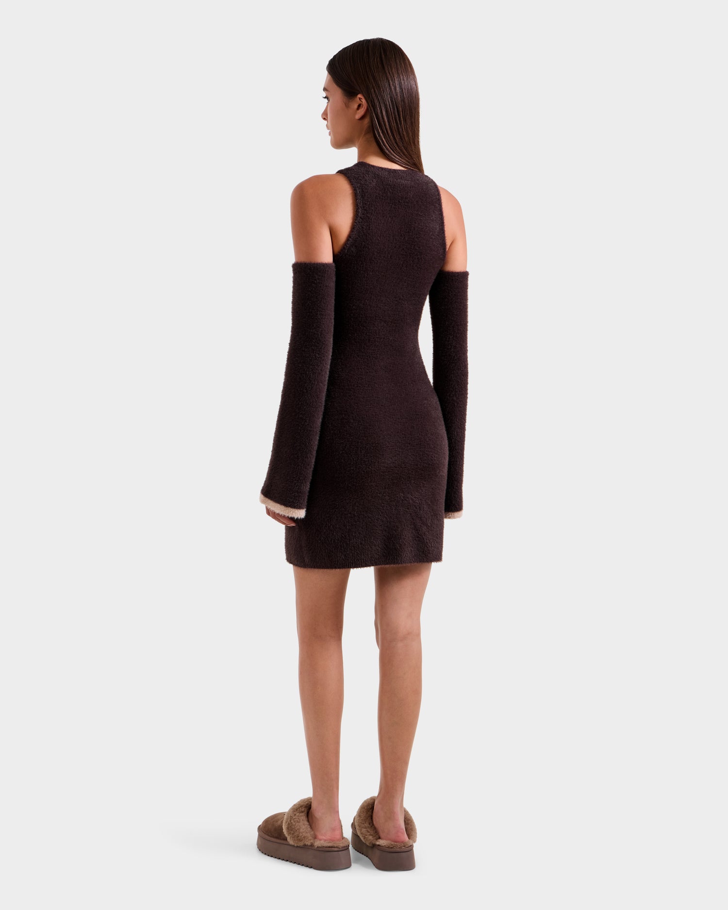 A woman faces away, wearing the ZARAH - 8525696 Espresso sleeveless fuzzy knit dress by Rudsak with matching detached sleeves and brown slippers, embodying après-ski style against a plain light gray backdrop.