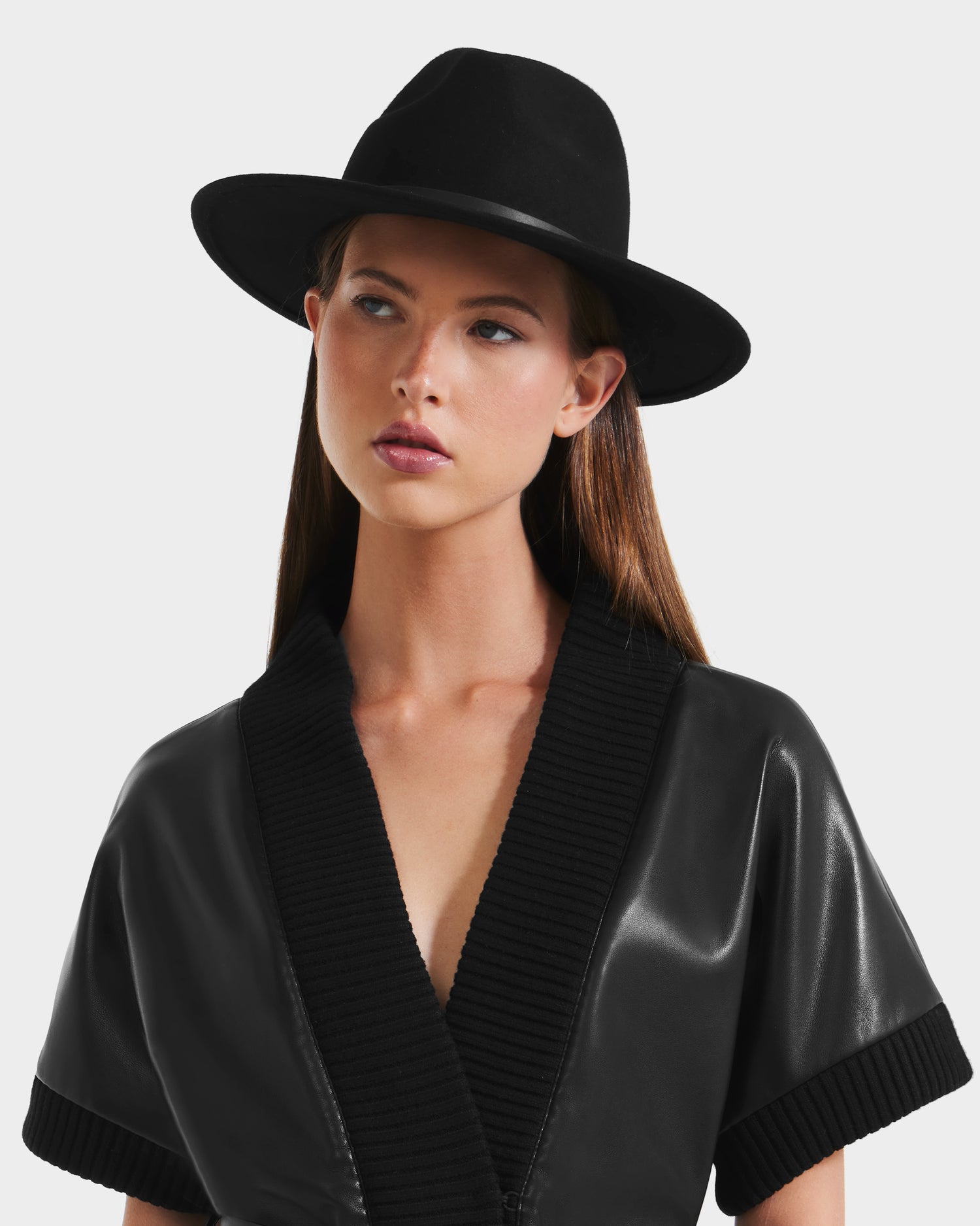 A woman wears the Rudsak ZELDA - 8425536 Black wool fedora and a black V-neck, short-sleeve faux leather top with ribbed trim as she looks to the side against a plain light background, adding elegance to her outfit.