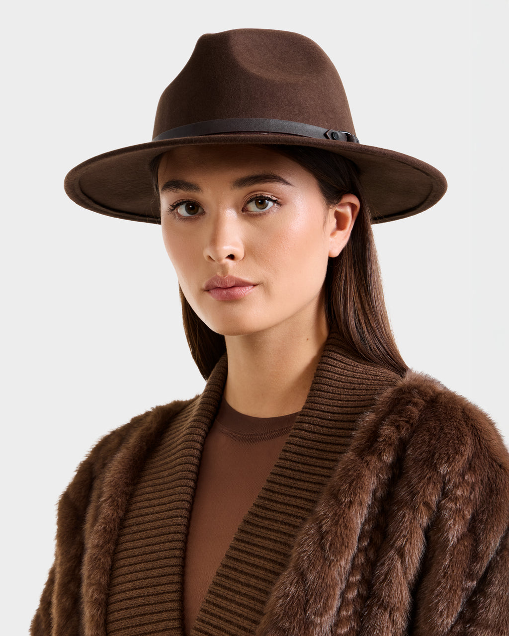 A woman wears the Rudsak ZELDA - 8425536 Espresso faux fur coat over a ribbed brown top and a wide-brim hat, looking directly at the camera against a plain light background.