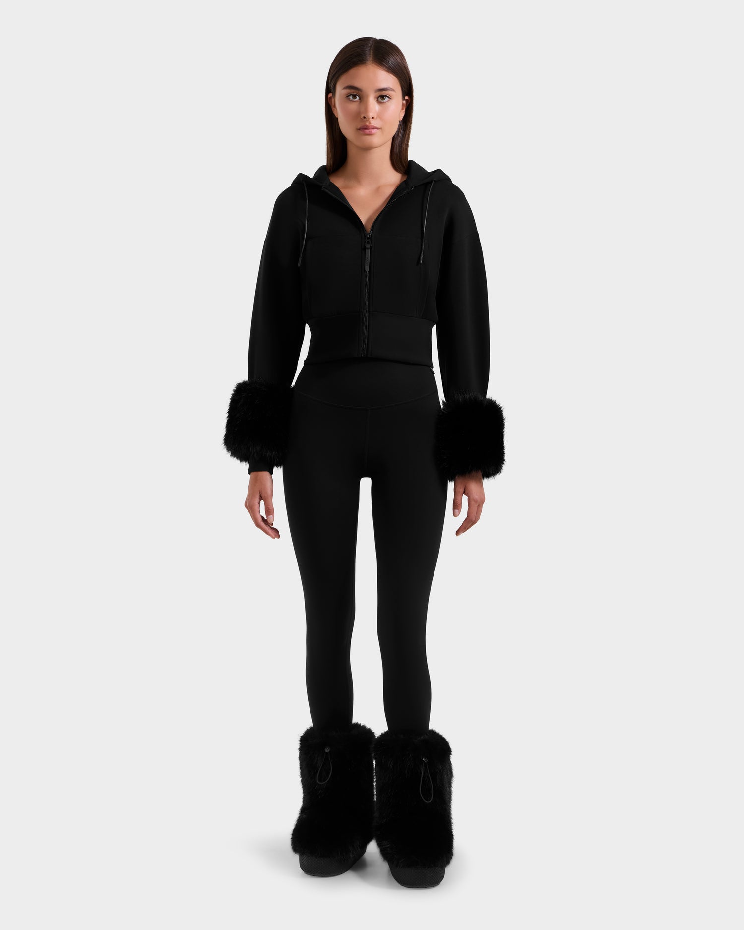 A woman faces forward in the ZILLIA - 8525673 Black cropped hoodie by Rudsak with faux fur cuffs, black leggings, and black fur boots, channeling après-ski chic against a plain light gray background.