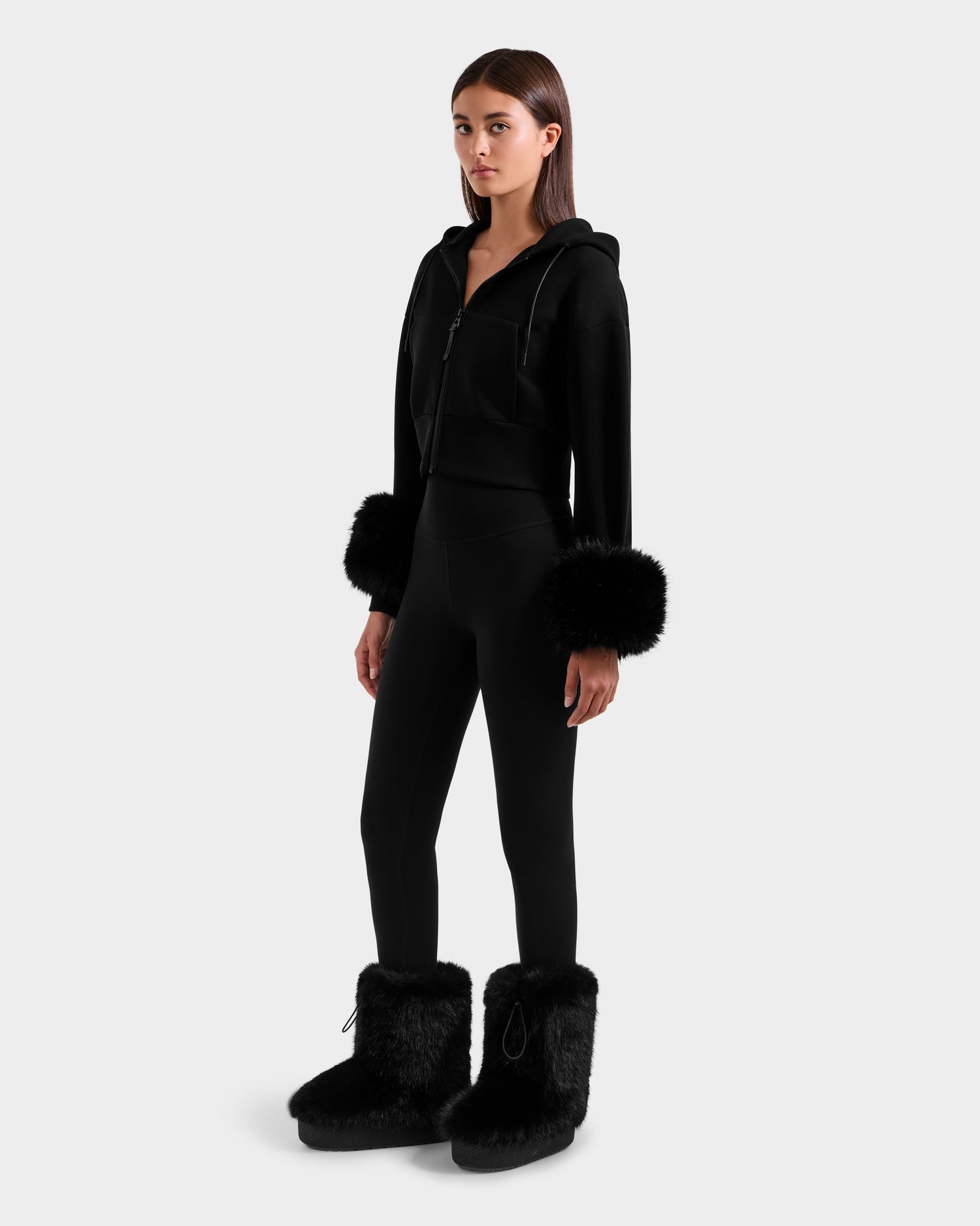 A woman faces forward in the Rudsak ZILLIA - 8525673 Black cropped hoodie with faux fur cuffs, paired with matching black leggings and furry boots, embodying après-ski chic against a plain light gray background.