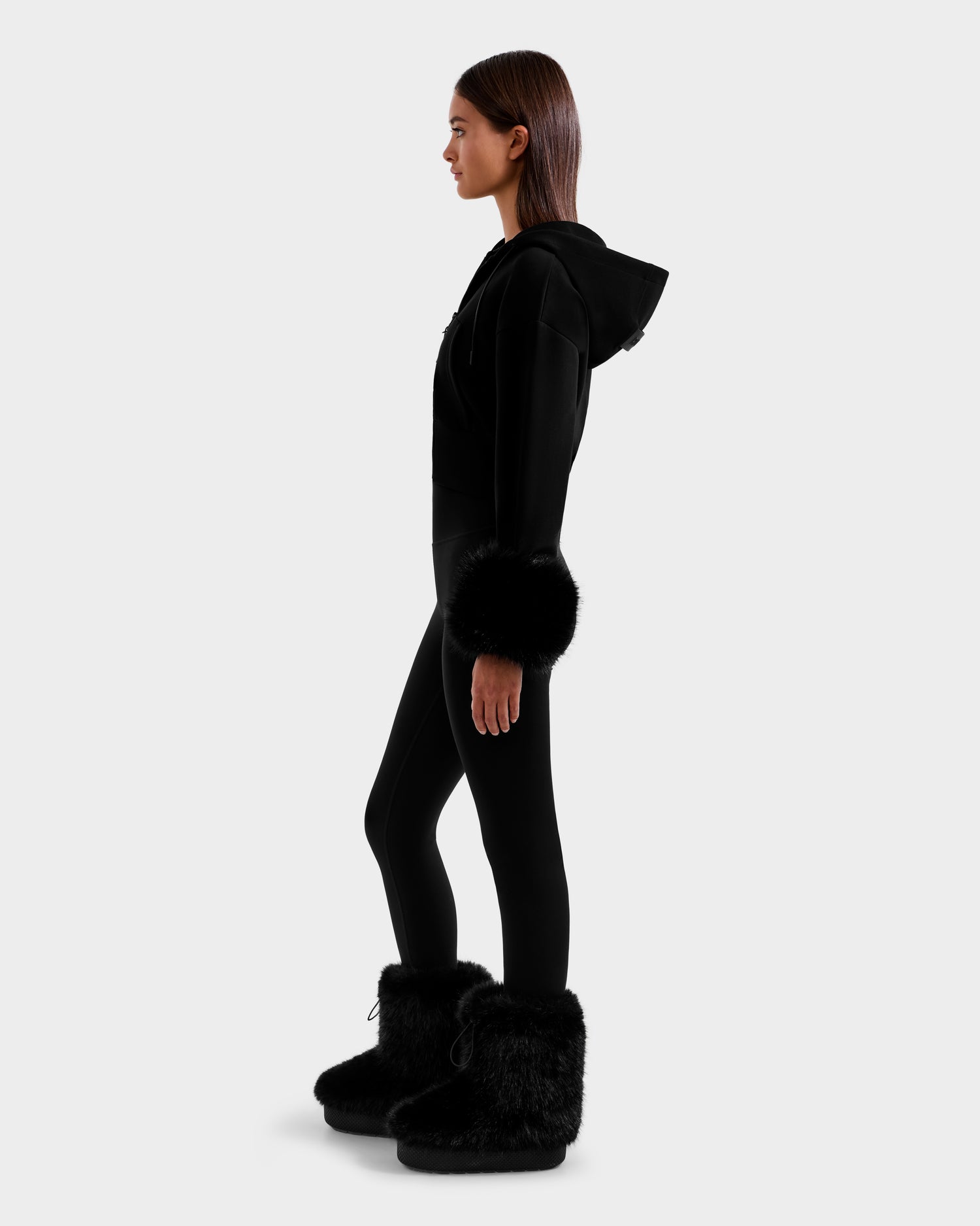 A woman in profile wears the Rudsak ZILLIA - 8525673 Black ensemble—an all-black après-ski look with a cropped hoodie, black leggings, large faux fur cuffs, and furry boots—set against a plain light gray background.