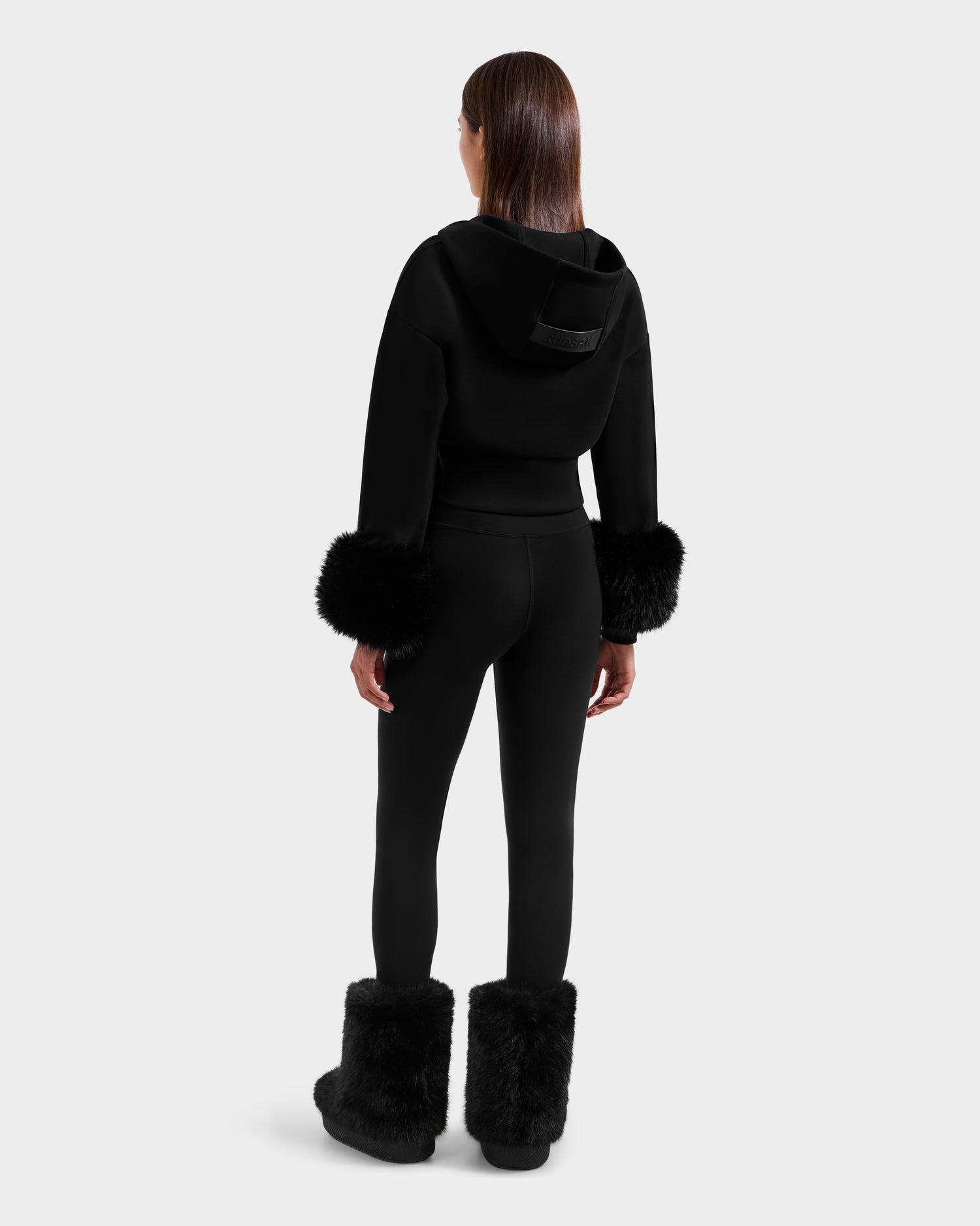 A woman faces away, wearing the Rudsak ZILLIA - 8525673 Black women’s cropped hoodie with faux fur wrist cuffs, black leggings, and furry boots—her straight brown hair down, exuding après-ski style on a plain light gray background.