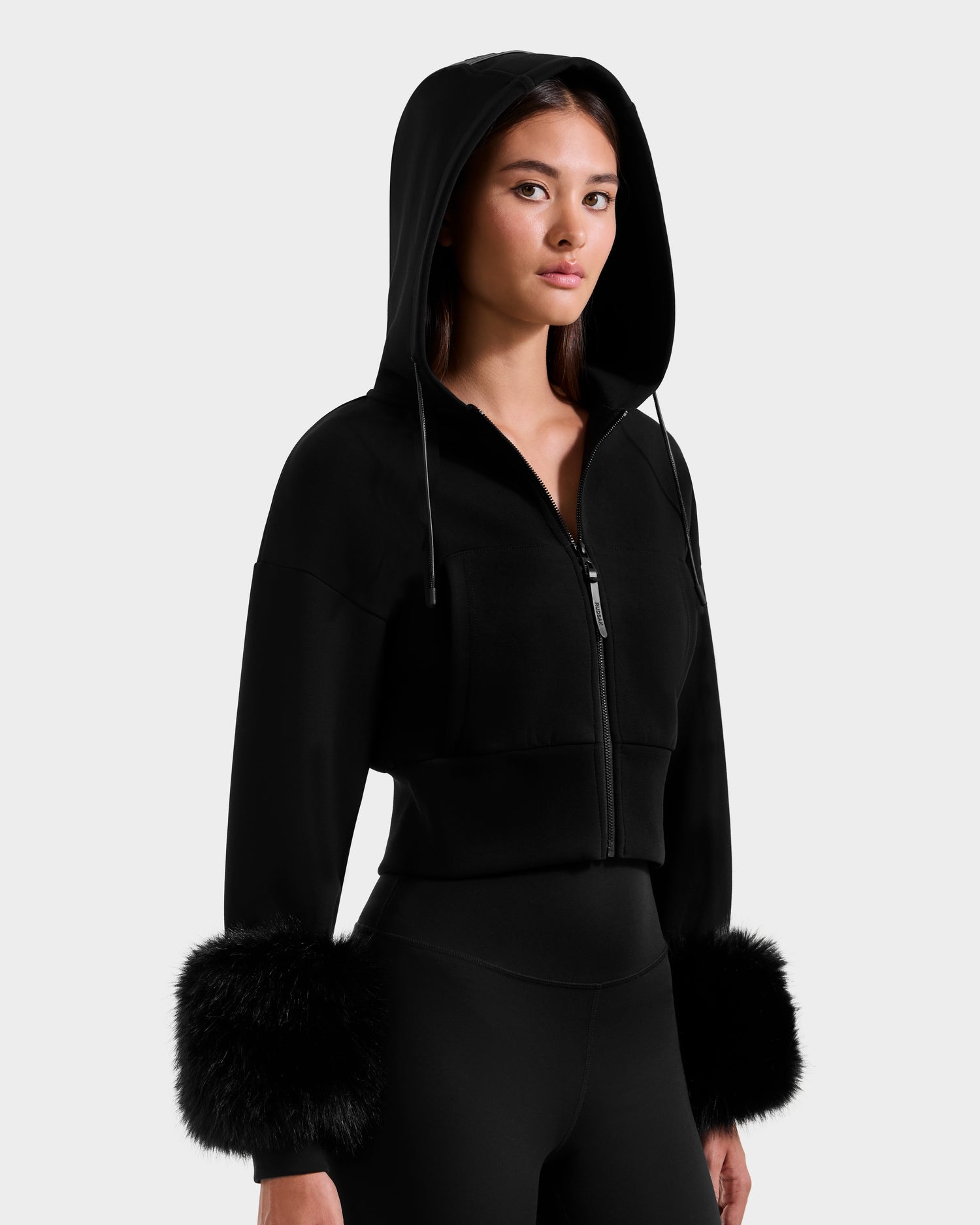 A woman models the Rudsak ZILLIA - 8525673 Black hooded zip-up jacket with faux fur cuffs and matching pants against a light gray background, exuding confidence at the camera.