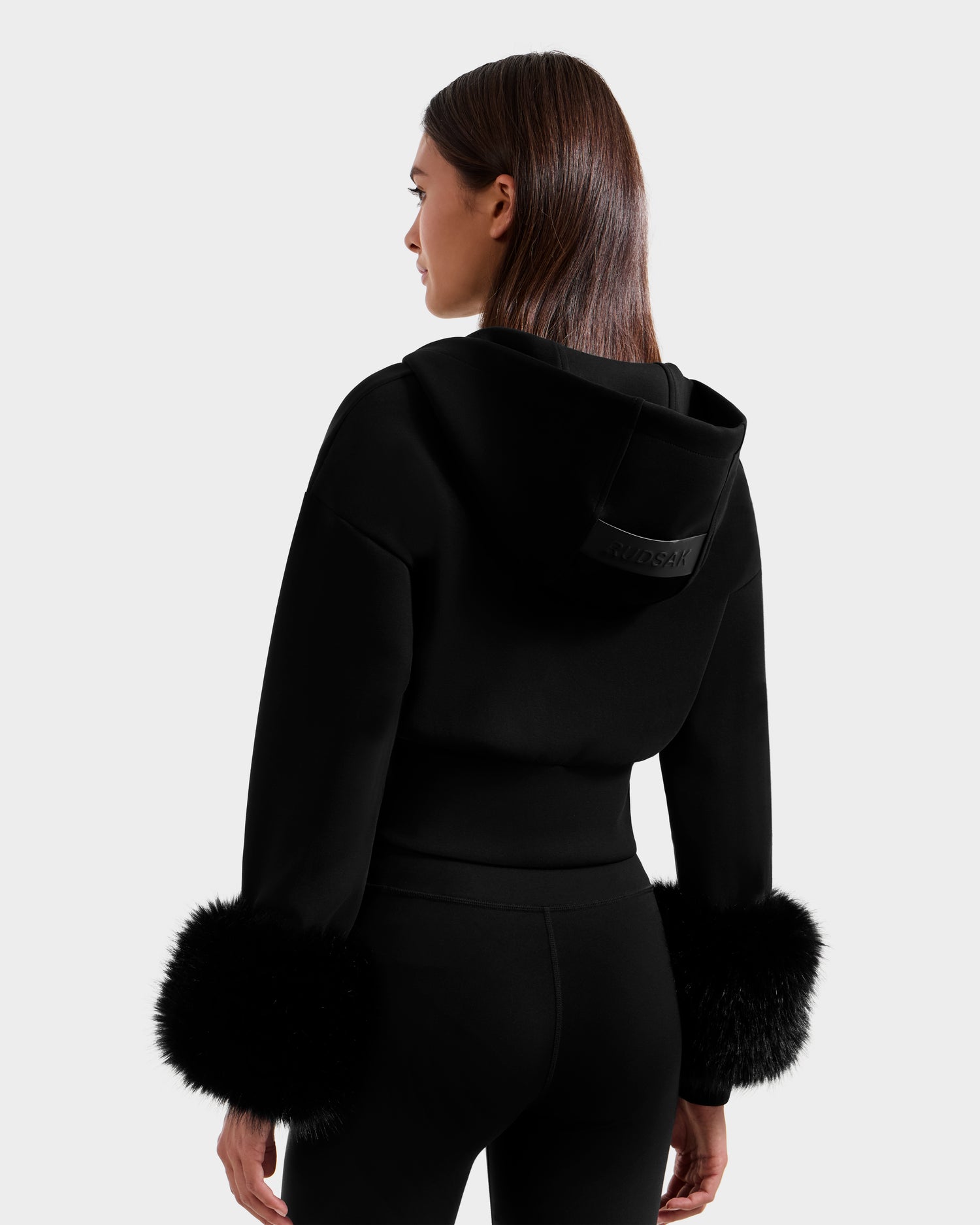 A woman with straight brown hair is seen from the back wearing the Rudsak ZILLIA - 8525673 Black women’s cropped hoodie with faux fur wrist cuffs and matching black pants, against a plain light gray background.