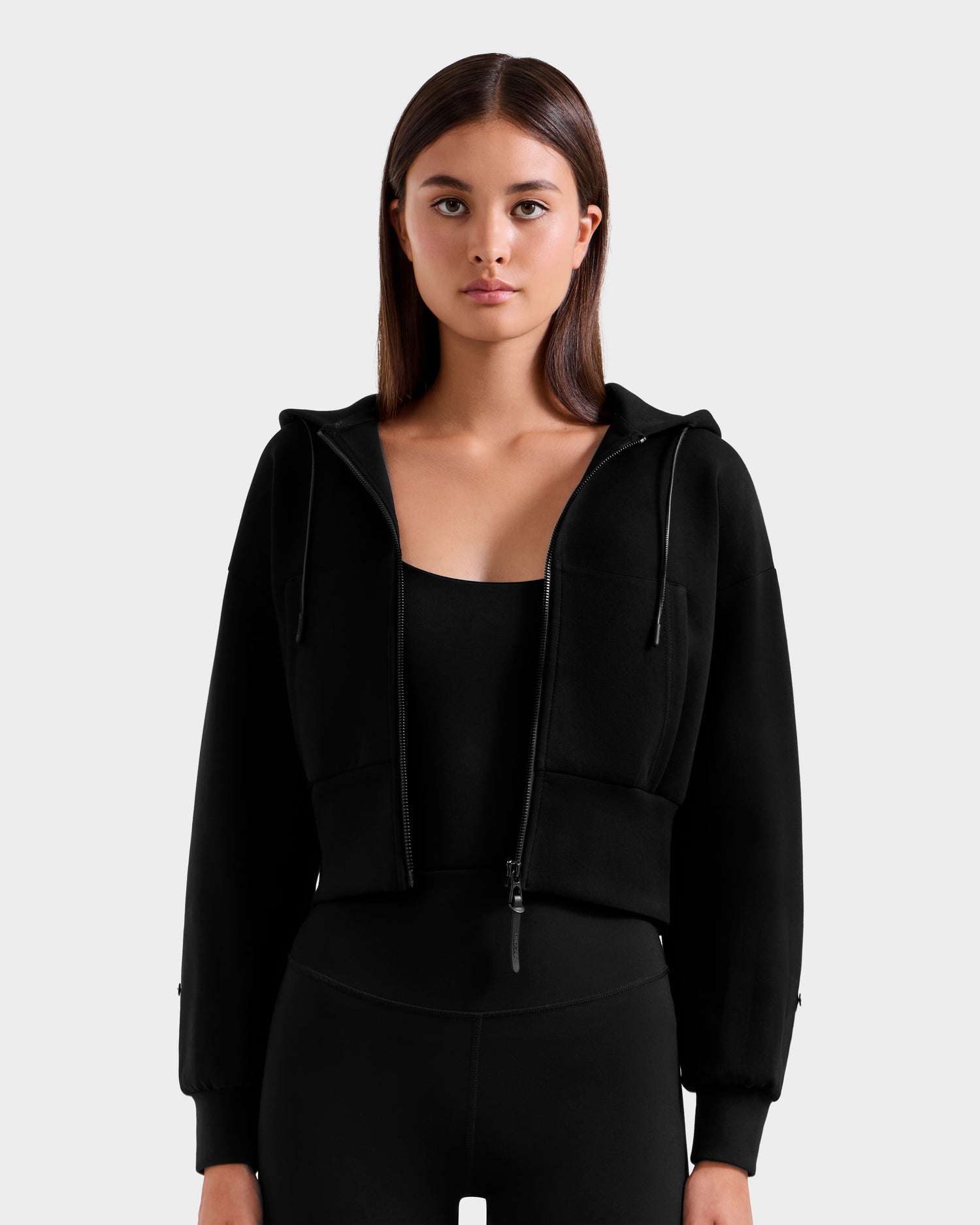 A woman with straight brown hair models the Rudsak ZILLIA - 8525673 Black cropped hoodie with faux fur wrist cuffs over black leggings, standing against a light gray background and looking at the camera with a neutral, après-ski chic vibe.