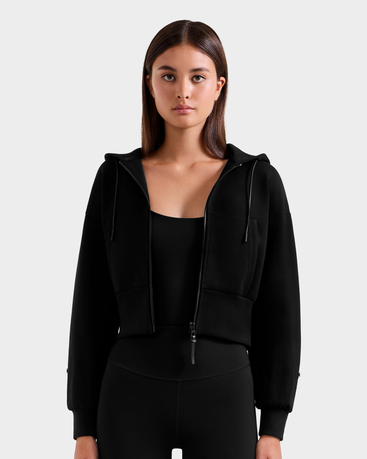 A woman with straight brown hair models the Rudsak ZILLIA - 8525673 Black cropped hoodie with faux fur wrist cuffs over black leggings, standing against a light gray background and looking at the camera with a neutral, après-ski chic vibe.