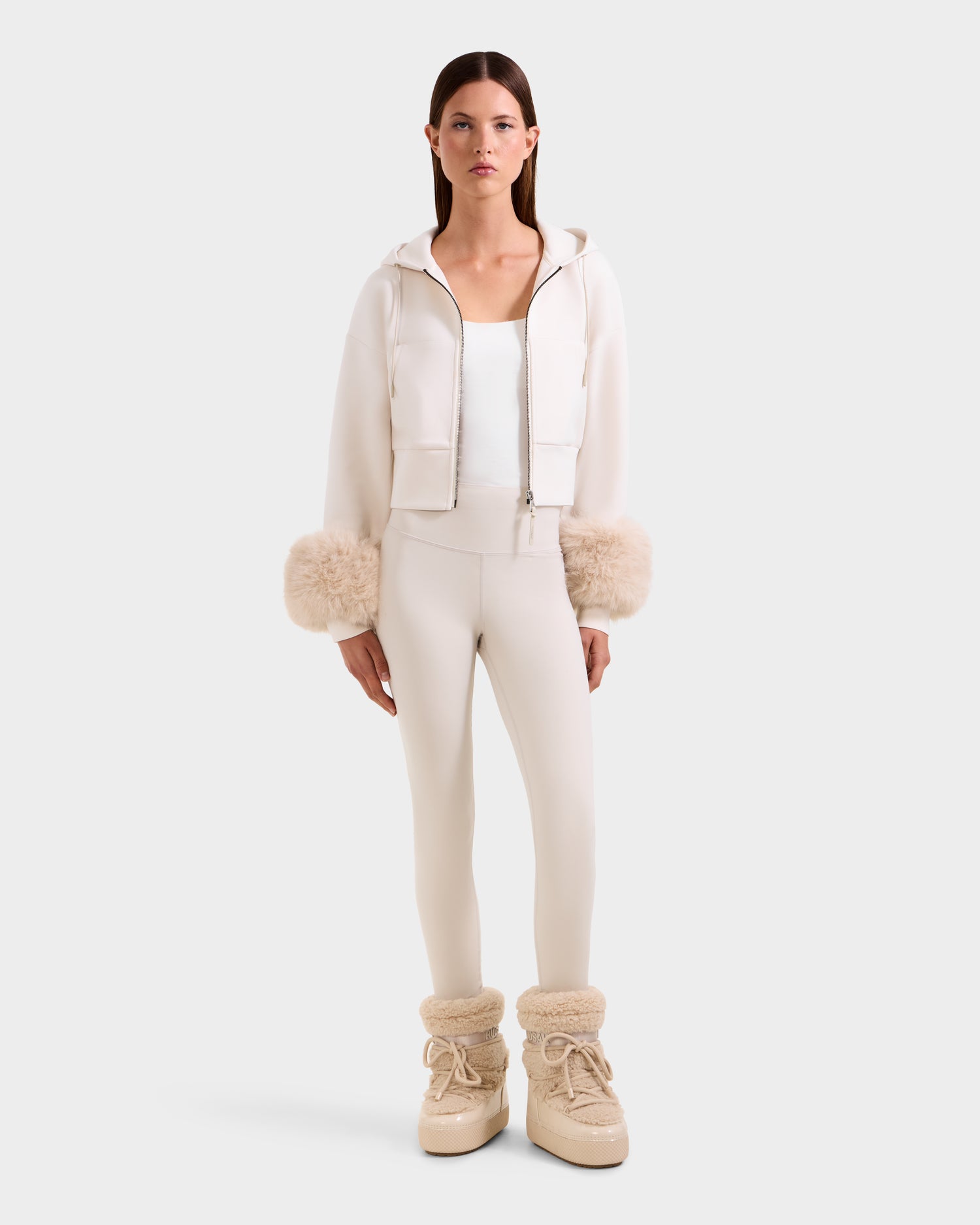 A woman faces forward wearing the Rudsak ZILLIA - 8525673 Vanilla cream cropped zip-up hoodie with faux fur cuffs, matching fitted pants, white top, and beige furry boots, set against a plain white background for a modern streetwear look.