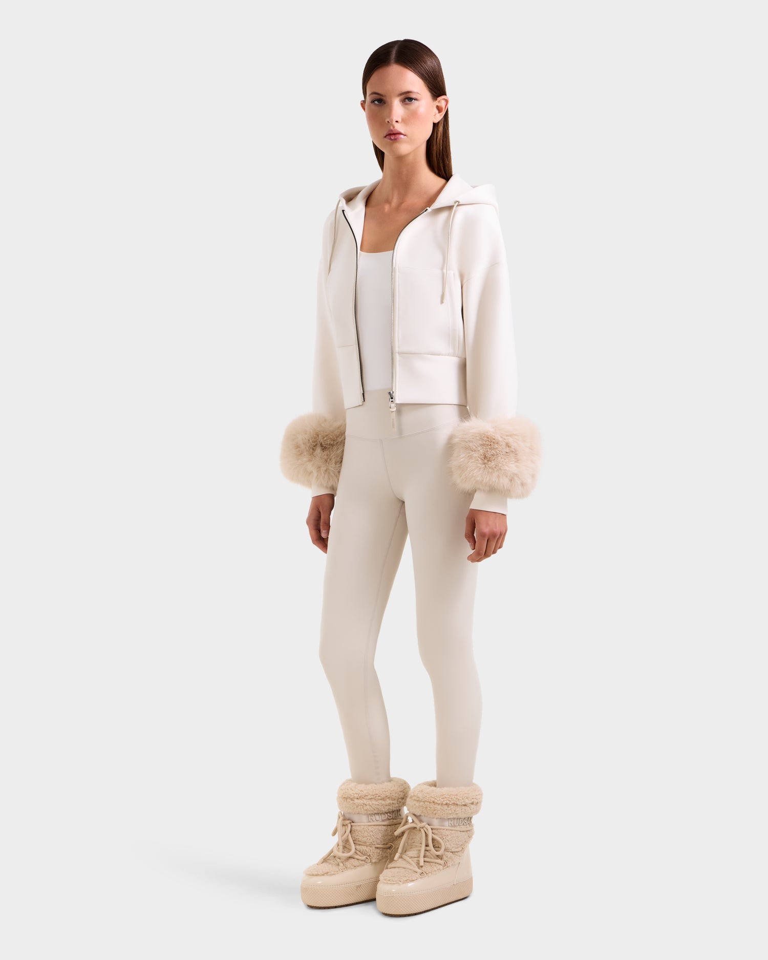 A woman stands against a plain light background wearing the Rudsak ZILLIA - 8525673 Vanilla, a white cropped zip-up hoodie with faux fur wrist cuffs—an elevated take on women's streetwear.