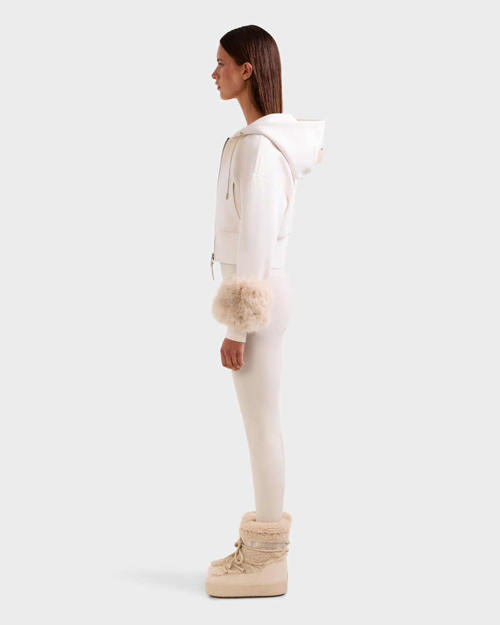 A woman stands in profile wearing the Rudsak ZILLIA - 8525673 Vanilla cropped zip-up hoodie with faux fur wrist cuffs, matching leggings, and beige fur-trimmed boots, creating a chic women's streetwear look against a plain light background.