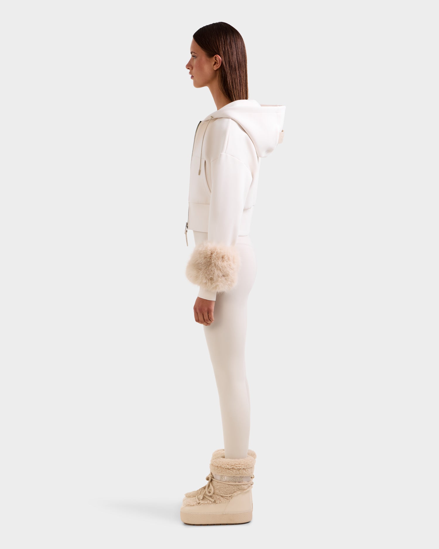 A woman stands in profile wearing the Rudsak ZILLIA - 8525673 Vanilla cropped zip-up hoodie with faux fur wrist cuffs, matching leggings, and beige fur-trimmed boots, creating a chic women's streetwear look against a plain light background.