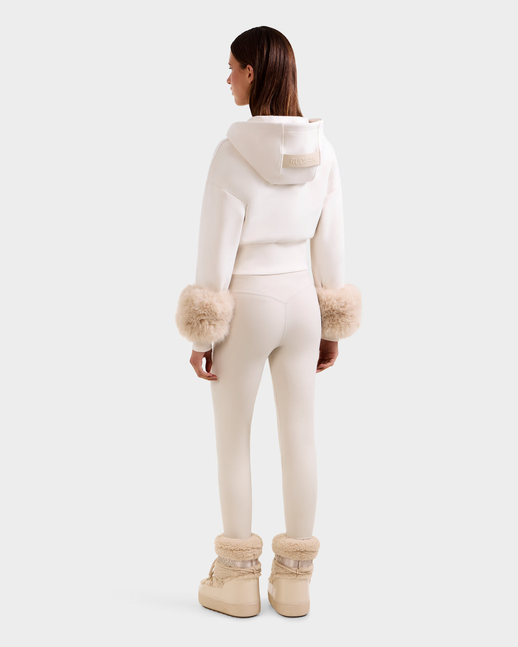 A woman with straight brown hair faces away, wearing the Rudsak ZILLIA - 8525673 Vanilla cropped zip-up hoodie with faux fur cuffs, matching leggings, and beige fur-trimmed boots against a plain white background.