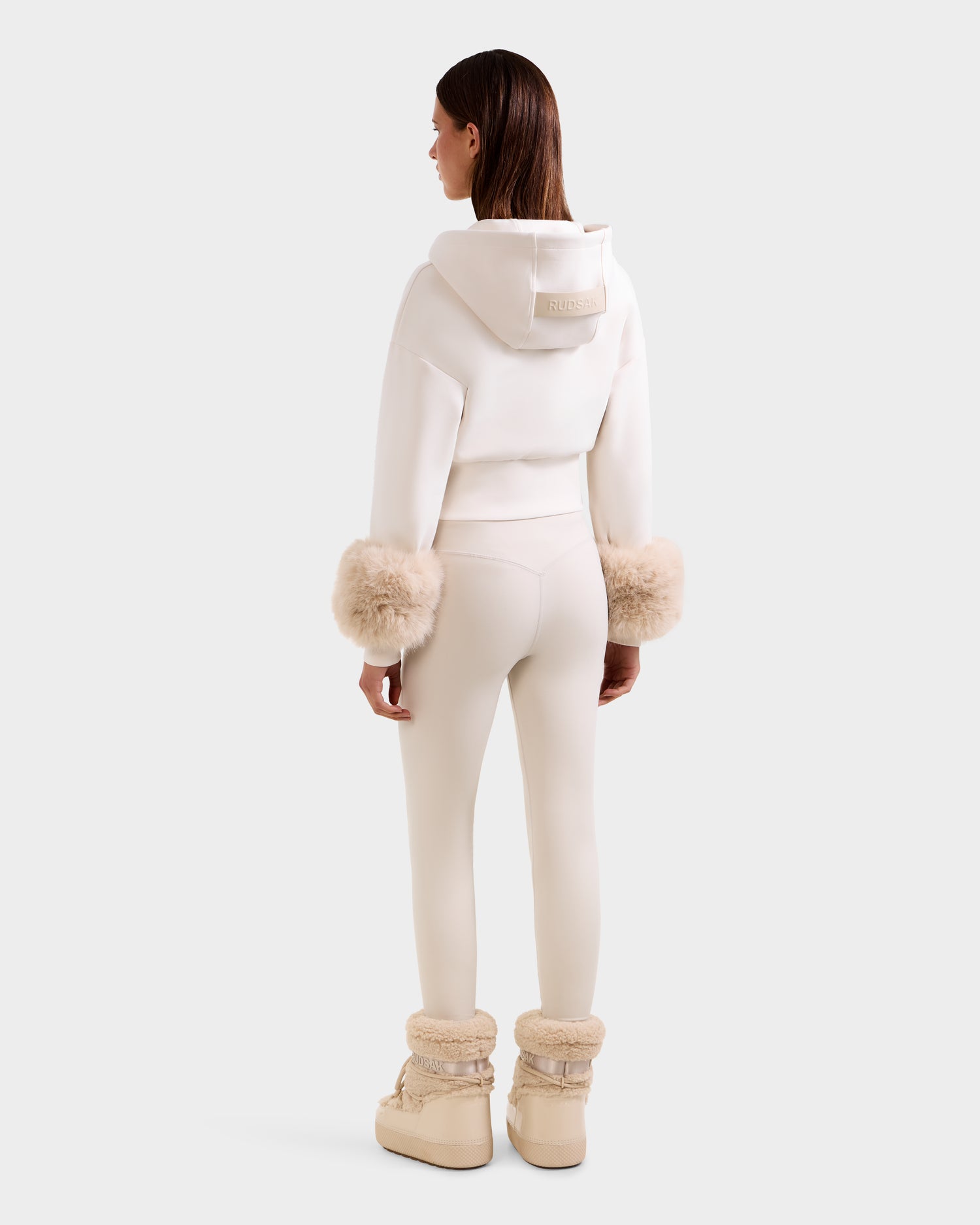 A woman with straight brown hair faces away, wearing the Rudsak ZILLIA - 8525673 Vanilla cropped zip-up hoodie with faux fur cuffs, matching leggings, and beige fur-trimmed boots against a plain white background.