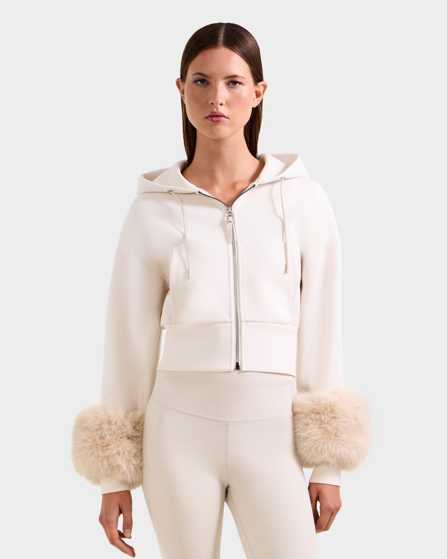 A woman models the Rudsak ZILLIA - 8525673 Vanilla: a cream cropped zip-up hoodie with oversized faux fur cuffs and matching high-waisted fitted pants, set against a simple backdrop—chic streetwear style.