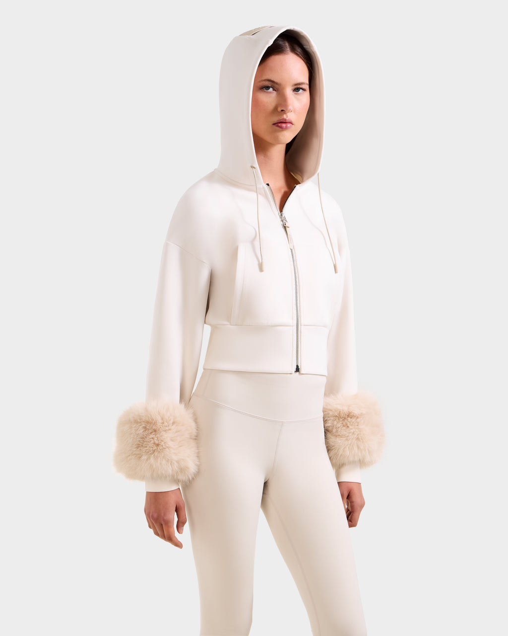 A woman models the ZILLIA - 8525673 Vanilla by Rudsak: a cream cropped zip-up hoodie with faux fur wrist cuffs and matching high-waisted pants, embodying chic women's streetwear against a plain background.