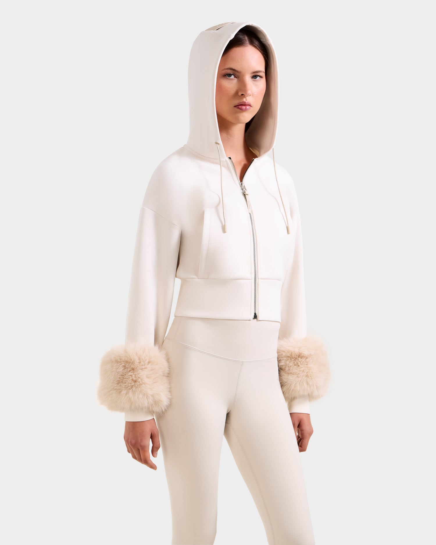 A woman models the ZILLIA - 8525673 Vanilla by Rudsak: a cream cropped zip-up hoodie with faux fur wrist cuffs and matching high-waisted pants, embodying chic women's streetwear against a plain background.