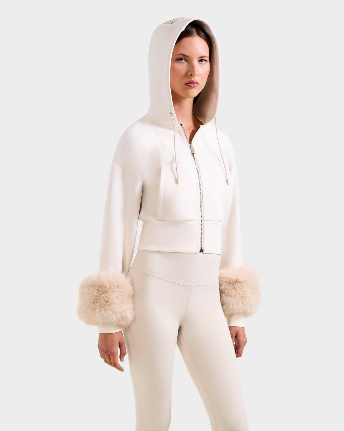A woman models the ZILLIA - 8525673 Vanilla by Rudsak: a cream cropped zip-up hoodie with faux fur wrist cuffs and matching high-waisted pants, embodying chic women's streetwear against a plain background.