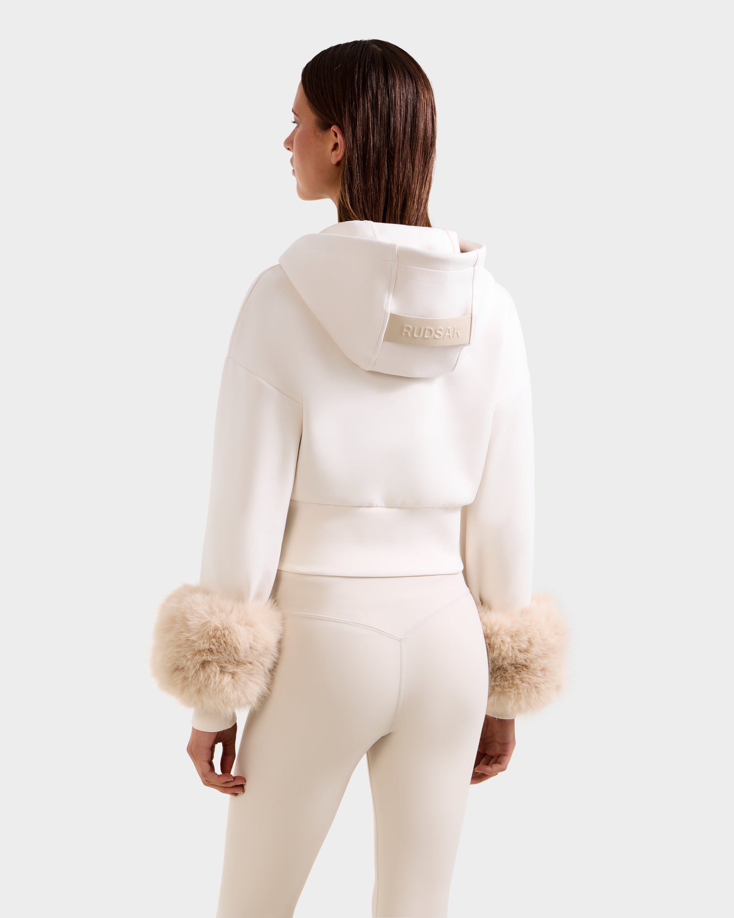 A woman models the Rudsak ZILLIA - 8525673 Vanilla set, featuring a cream-colored cropped zip-up hoodie with faux fur cuffs and matching fitted pants, showcasing "RUDSAK" embroidered on the back of the hood.