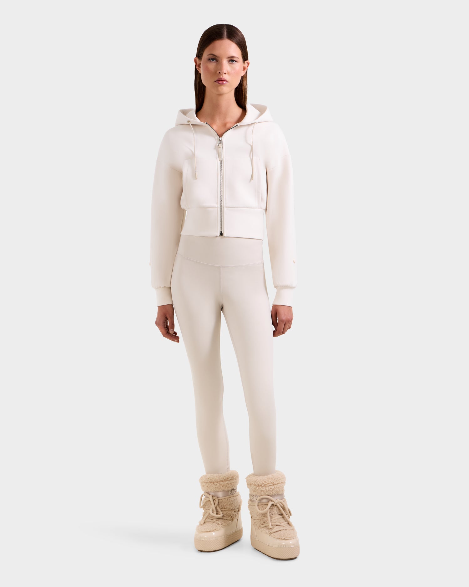 A woman with straight brown hair and a neutral expression showcases effortless streetwear in Rudsak's ZILLIA - 8525673 Vanilla: a cropped zip-up hoodie, matching high-waisted leggings, and light beige, fluffy boots against a plain background.