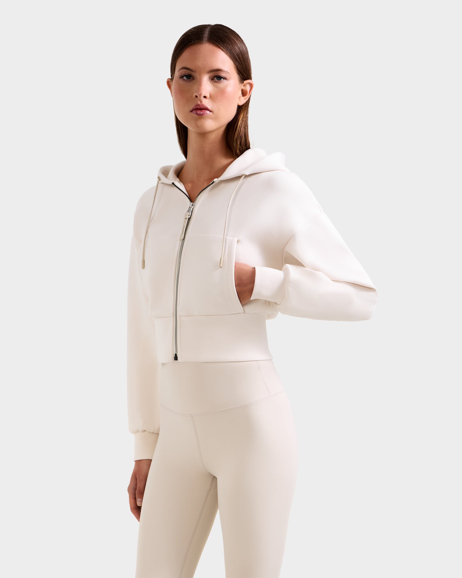 A woman models Rudsak’s ZILLIA - 8525673 Vanilla: a cream cropped zip-up hoodie with faux fur cuffs and matching high-waisted leggings, standing against a light gray background and showcasing modern women’s streetwear style.