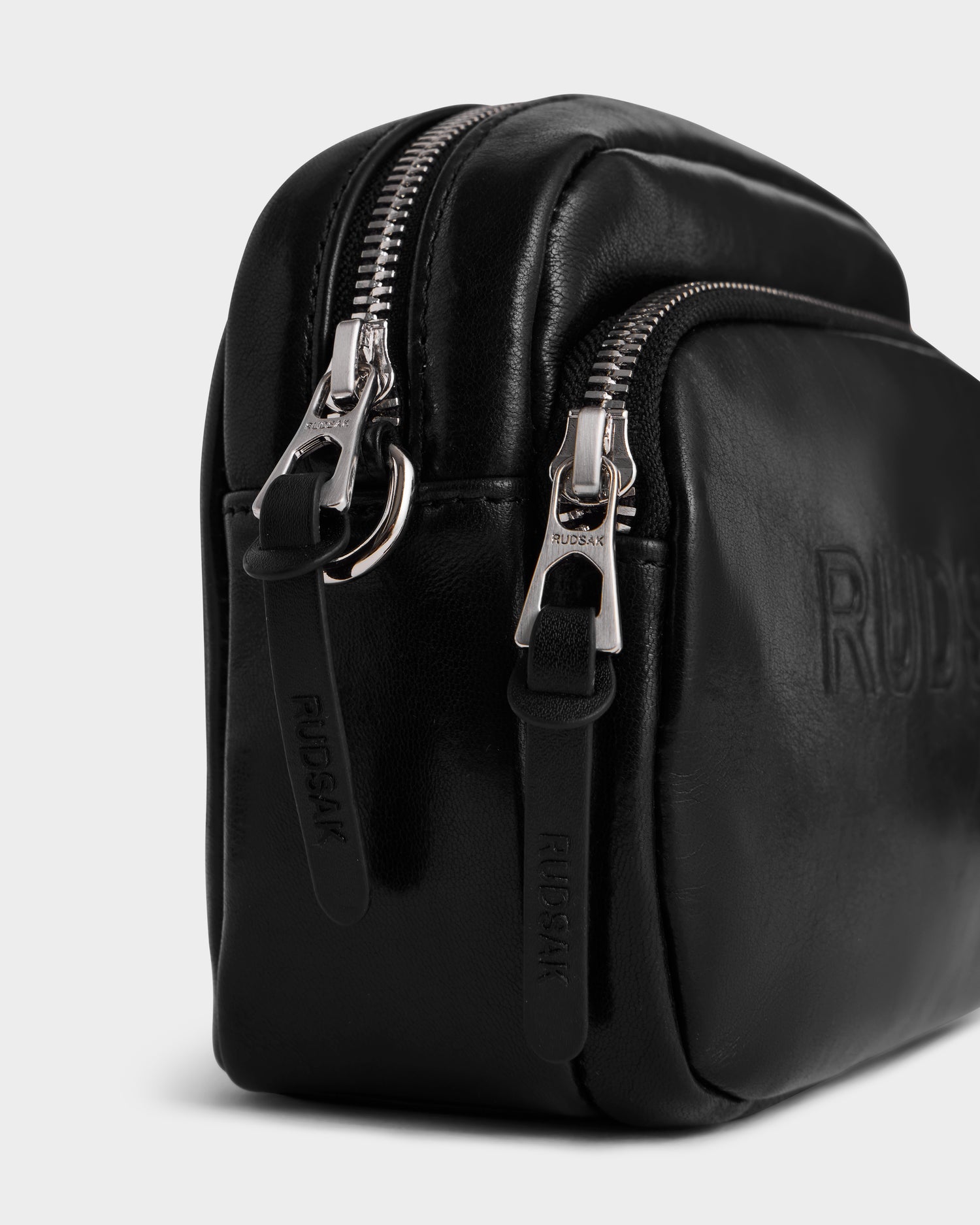 Close-up of the Rudsak ZOYA L - 8325543 Black crossbody bag with smooth leather, embossed logo, two silver zippers with leather pulls, adjustable strap, and a front zip pocket. Sleek and modern look against a plain background.