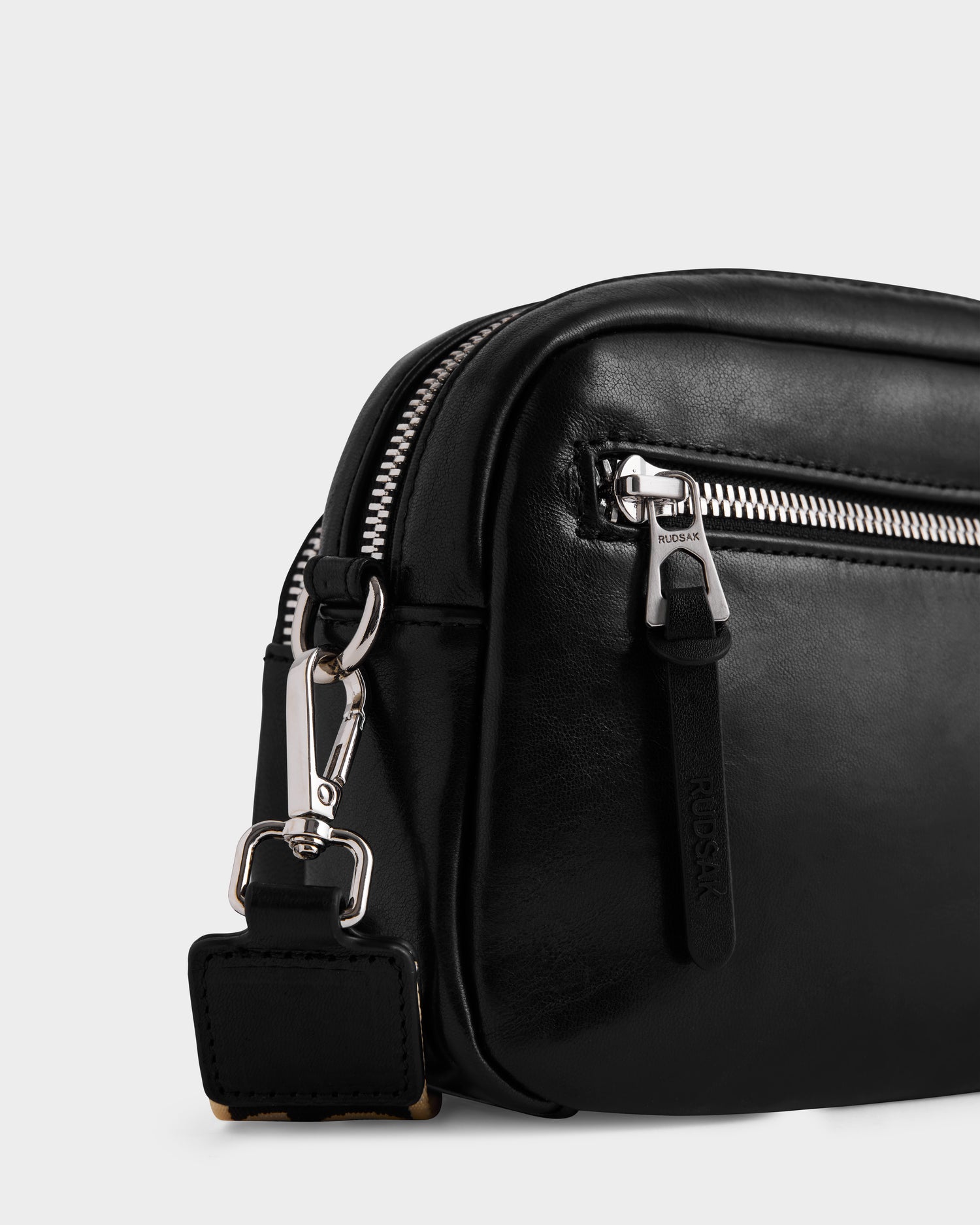 Close-up of the Rudsak ZOYA L - 8325543 Black crossbody bag featuring smooth black leather, a front pocket with silver zipper and branded pull, plus an adjustable detachable strap with a silver clasp. Plain white background.