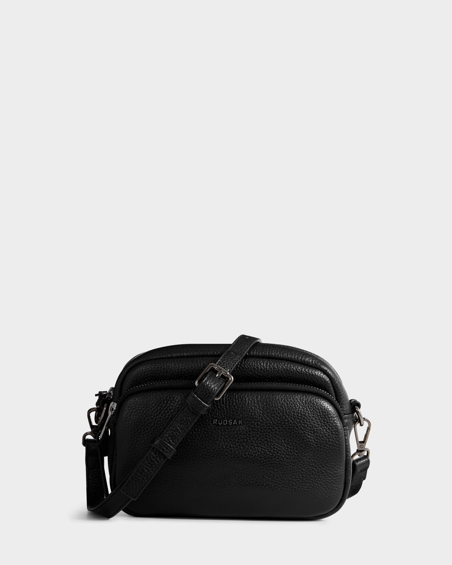The Rudsak ZOYA - L325015 Black is a small women’s crossbody bag in pebbled leather with an adjustable strap, silver hardware, and two zippered compartments, shown on a plain light gray background.