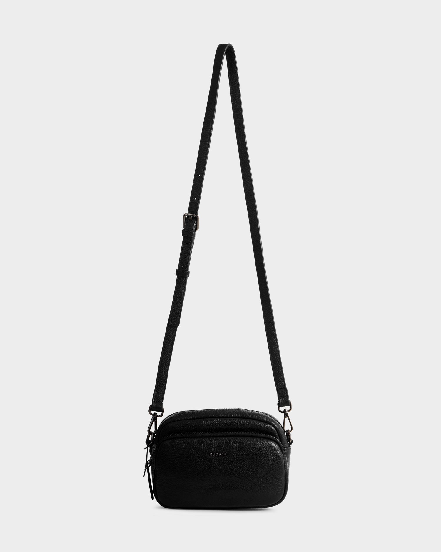 The Rudsak ZOYA - L325015 Black is a small women’s crossbody bag in pebbled leather, featuring an adjustable strap, two zippered compartments, and a minimal design, shown against a light gray background.