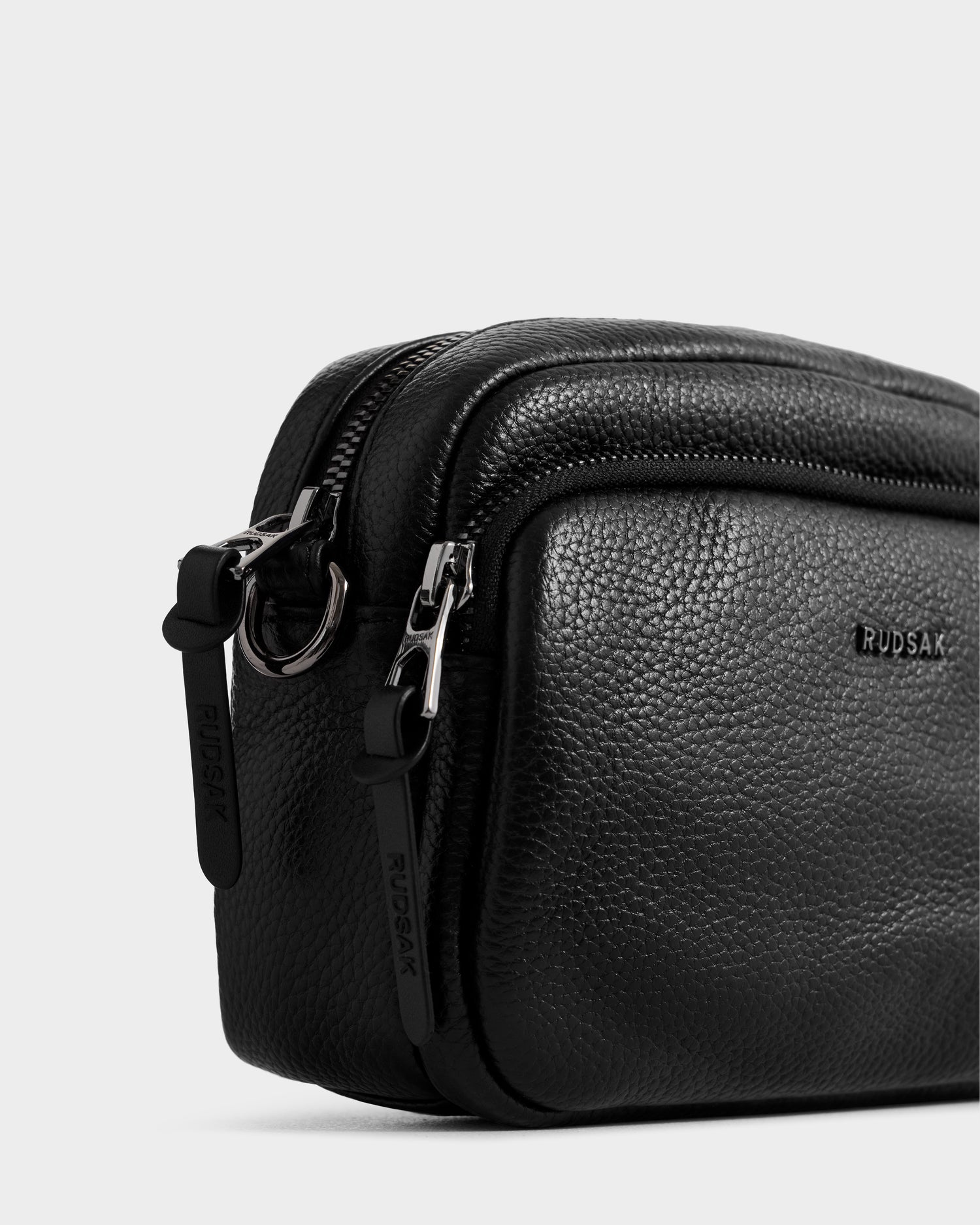Close-up of the Rudsak ZOYA - L325015 Black women’s crossbody bag in black pebbled leather, featuring two zippered compartments, silver hardware, and “Rudsak” embossed on the front—an ideal everyday accessory.
