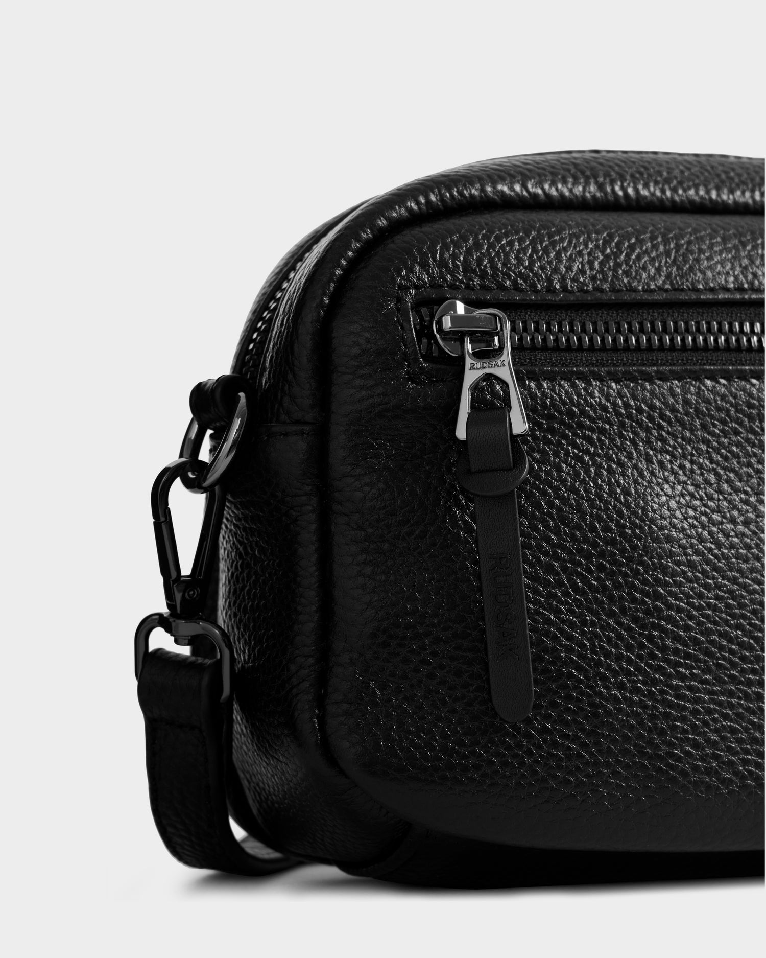 Close-up of the Rudsak ZOYA - L325015 Black women’s crossbody bag in black pebbled leather, featuring a zippered front pocket, metal zipper pull with strap, and an adjustable, detachable shoulder strap with metal clasp.