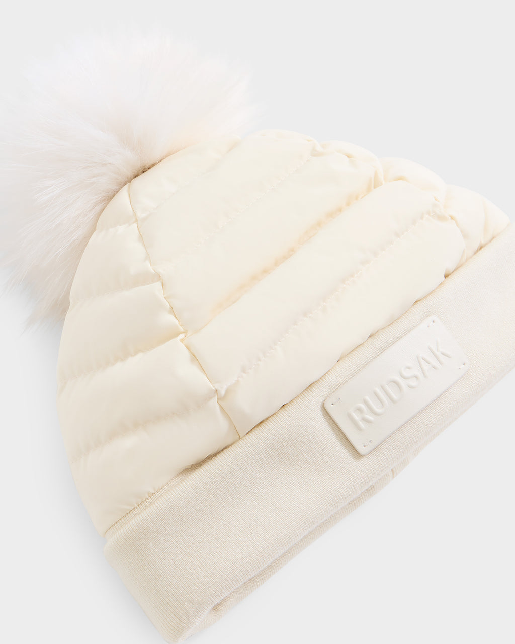 The GALA - 8424514 Iceland by Rudsak is a cream-colored, quilted women’s down puffer hat featuring a large removable white faux fur pom-pom on top, ribbed fabric at the brim, and a rectangular front patch labeled RUDSAK.