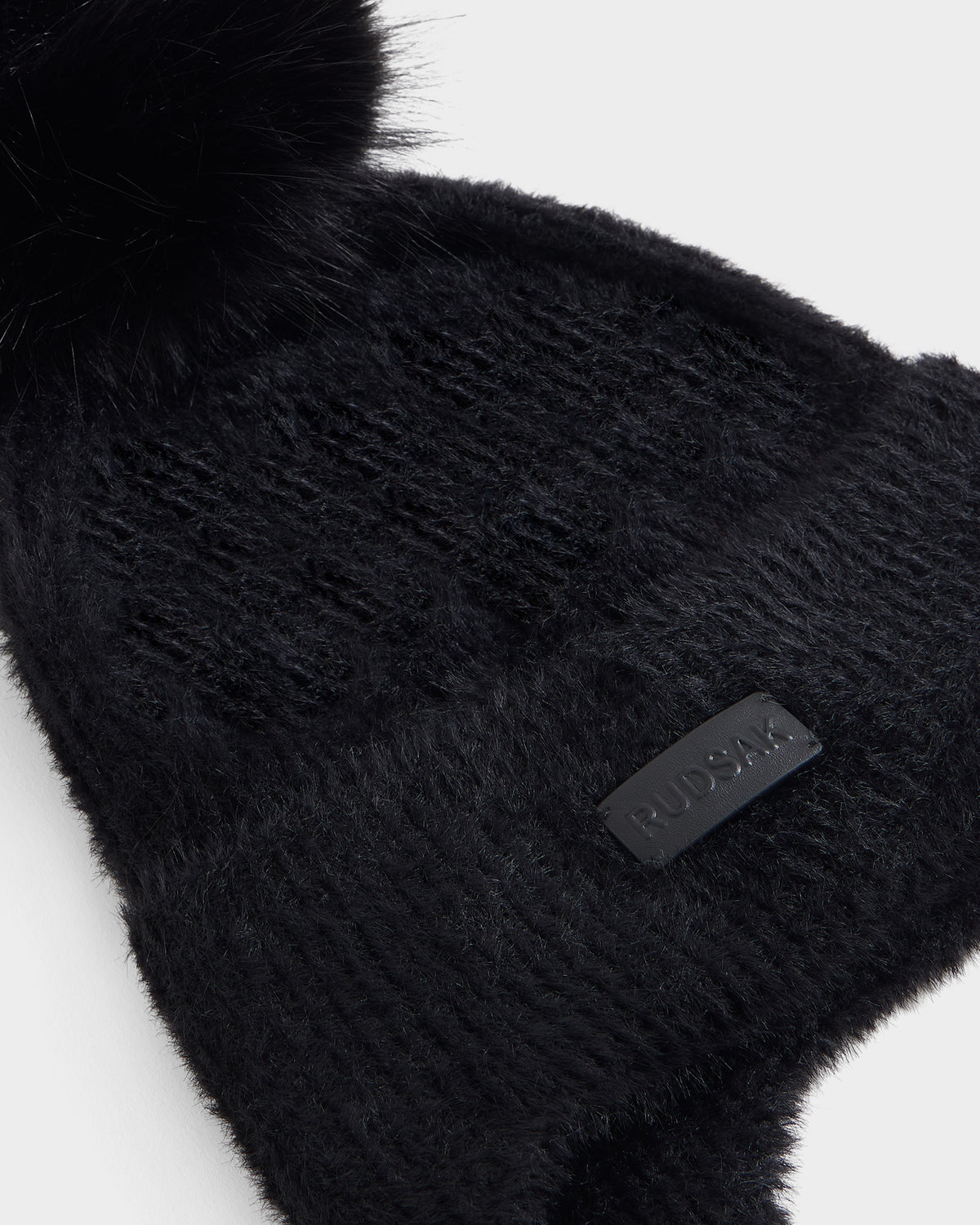 A close-up of the Rudsak ICE - 8426519 Black knit beanie, featuring a faux-fur pom-pom and a leather patch label, making it the perfect winter accessory for any après-ski hat collection.