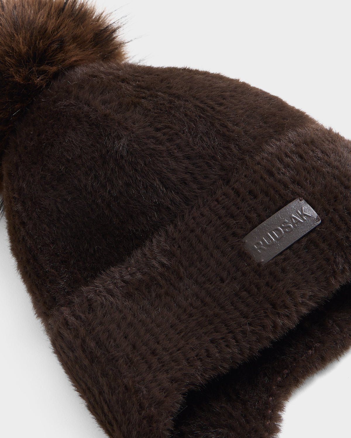 A close-up of the Rudsak ICE - 8426519 Espresso beanie, a dark brown, fuzzy winter accessory featuring a faux-fur pom-pom on top and a leather patch label that reads RUDSAK.
