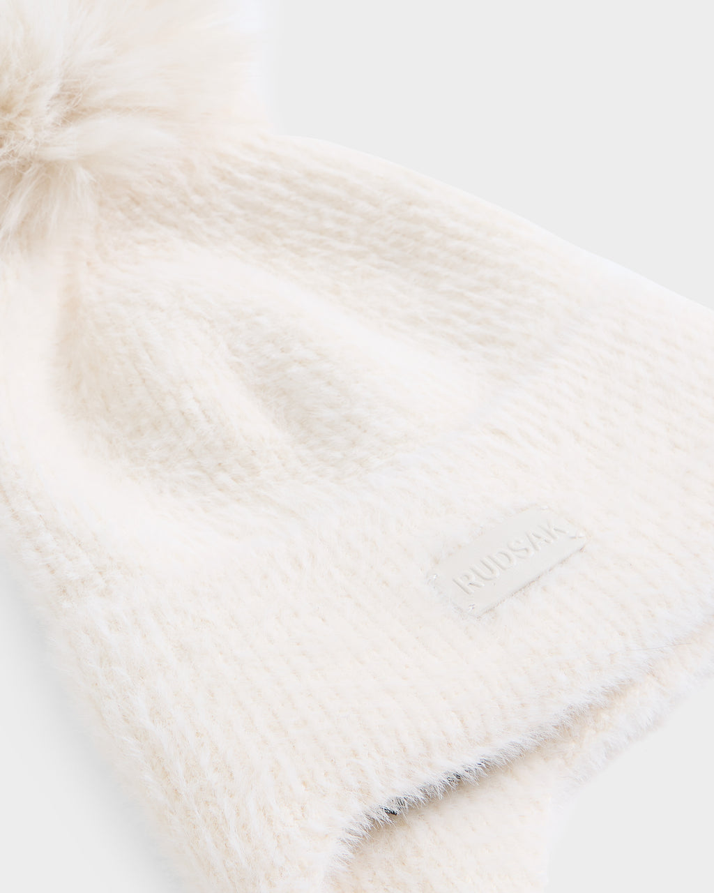 The Rudsak ICE - 8426519 Vanilla beanie features a cream-colored knit design topped with a fluffy faux-fur pom-pom. A small, rectangular patch with text is sewn onto the brim, and its soft, fuzzy texture makes it the perfect winter accessory. The product is photographed against a plain white background.