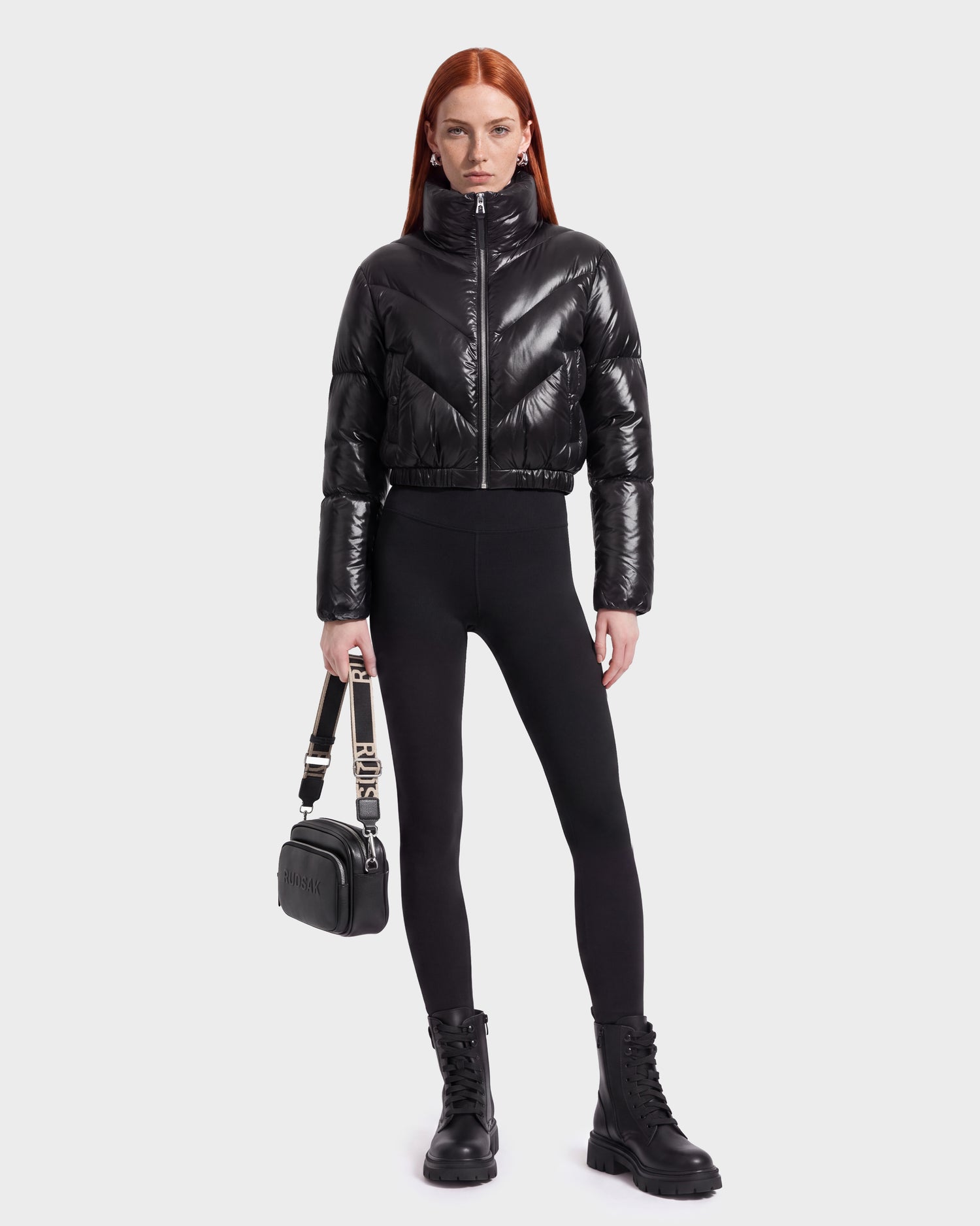 A person with long red hair wears the Rudsak ANSLIE C - 8126713 Black, a shiny, fashion-forward puffer jacket made with RDS-certified down, paired with black leggings, lace-up black boots, and holding a small black handbag against a plain light background.