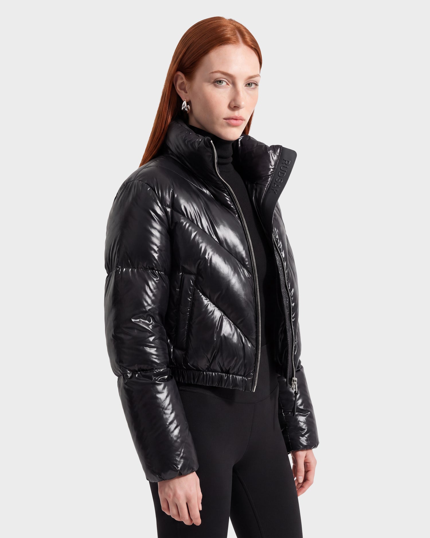 A woman with long red hair wears the Rudsak ANSLIE C - 8126713 Black, a shiny, fashion-forward women’s down puffer jacket, paired with a black turtleneck and black pants, standing against a plain light gray background.