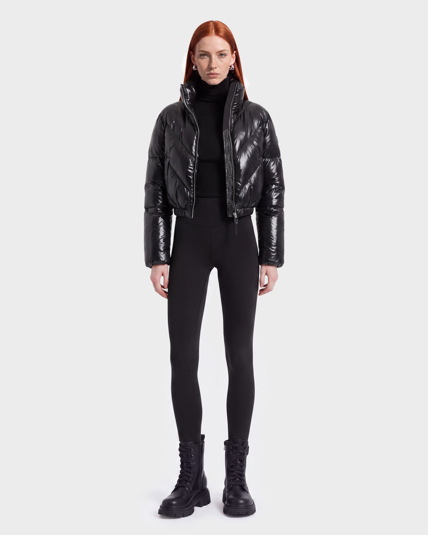 A person with long red hair wears the Rudsak ANSLIE C - 8126713 Black puffer jacket, paired with a black turtleneck, black leggings, and black lace-up boots, standing against a plain white background.
