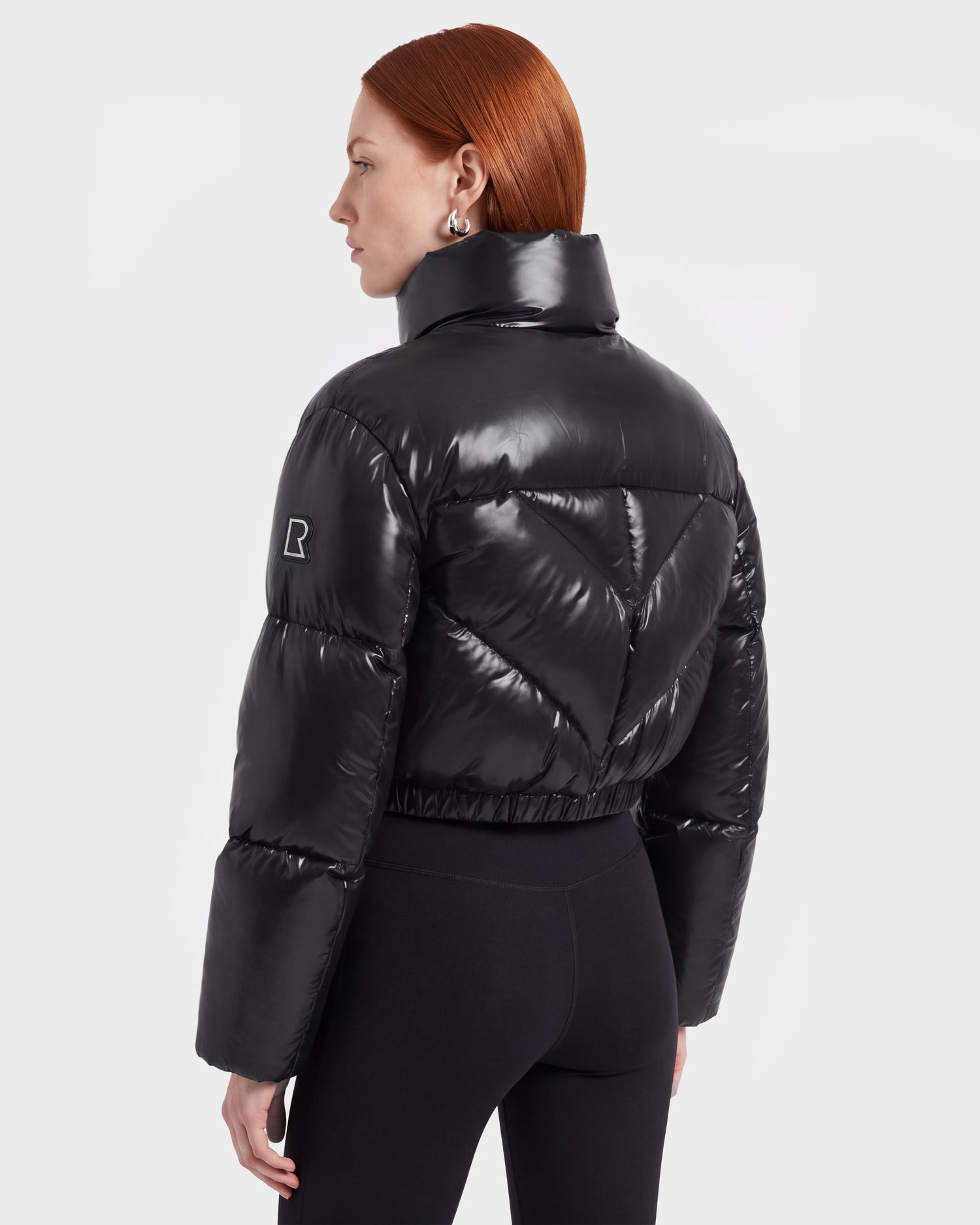 A woman with straight red hair is shown from the back wearing the fashion-forward Rudsak ANSLIE C - 8126713 Black women’s down puffer jacket, which features a shiny black finish, a high collar, a stitched V pattern, and an R on the upper sleeve. She pairs it with black pants.