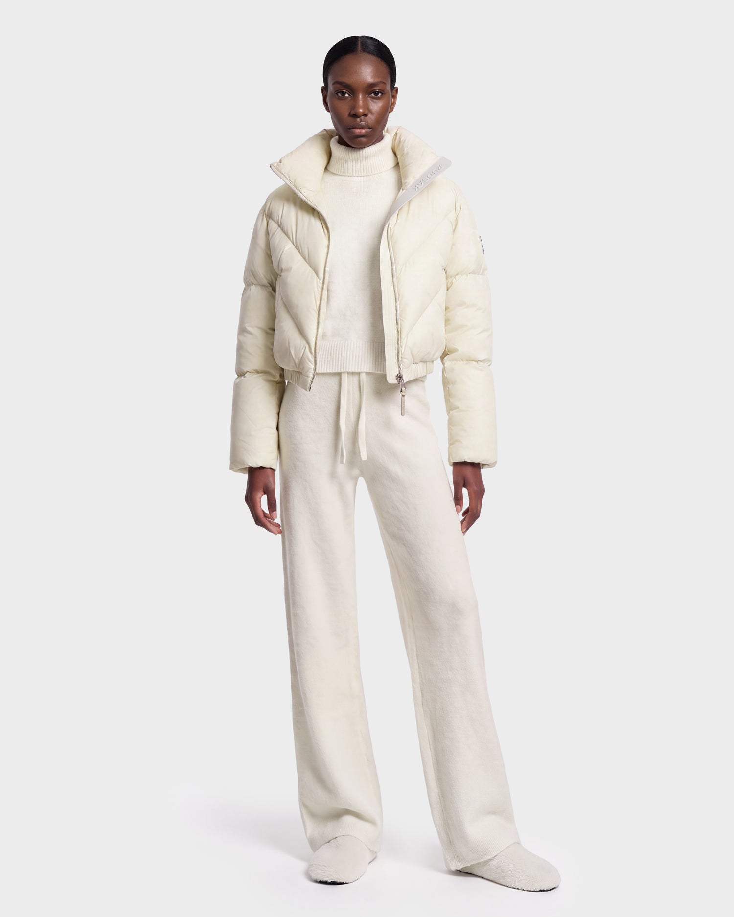 A person stands facing forward against a plain white background, wearing the Rudsak ANSLIE - 8126986 Vanilla off-white cropped puffer jacket filled with RDS-certified down, paired with a turtleneck sweater, wide-leg pants, and slippers.