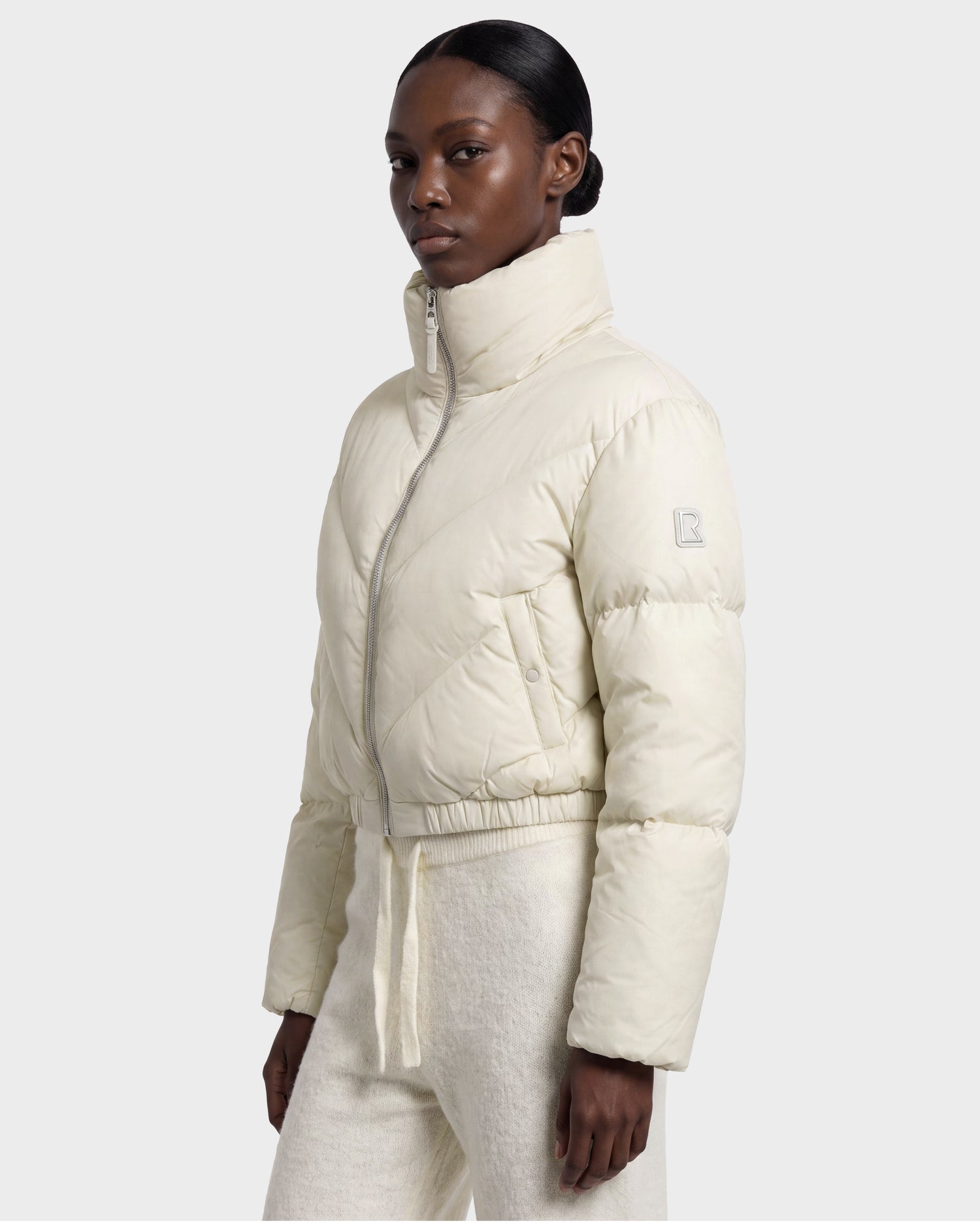 A woman wearing the Rudsak ANSLIE - 8126986 Vanilla light beige cropped puffer jacket with a high collar and matching pants stands against a plain background, looking slightly to the side.
