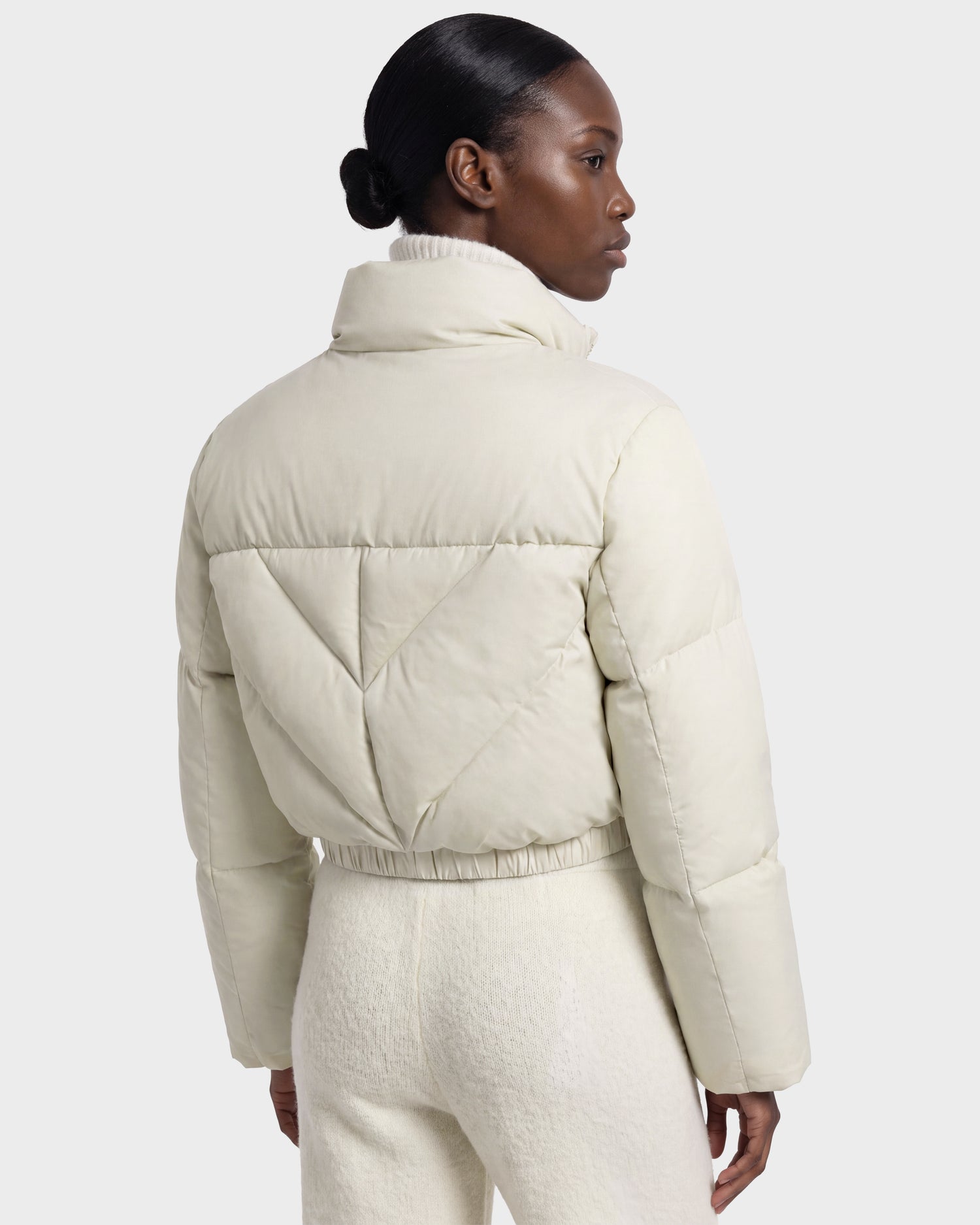 A person with dark hair in a low bun wears the Rudsak ANSLIE - 8126986 Vanilla, an RDS-certified down puffer jacket with matching pants, standing against a plain background and facing away from the camera.