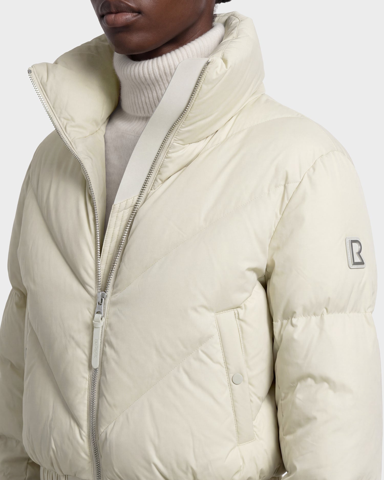 A person is wearing the Rudsak ANSLIE - 8126986 Vanilla, a cream-colored women’s cropped puffer jacket filled with RDS-certified down. This jacket features a zipper closure, high collar, side pockets, and a signature B logo patch on the sleeve, styled over a white turtleneck sweater.