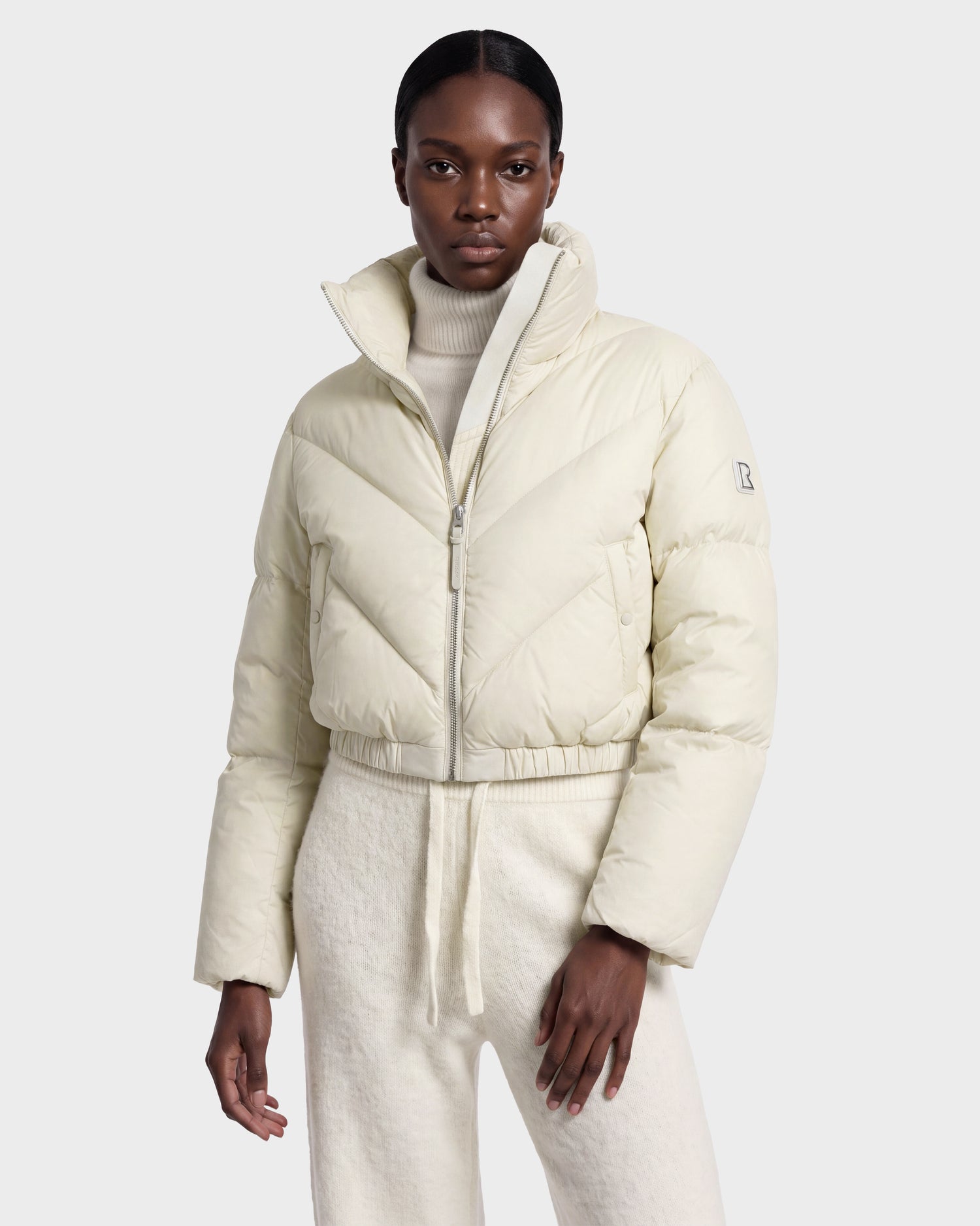 A person stands facing forward against a plain white background, dressed in the ANSLIE - 8126986 Vanilla cropped puffer jacket from Rudsak, layered over a matching turtleneck and pants. This women’s down puffer is filled with RDS-certified down, offering both warmth and sustainable comfort.