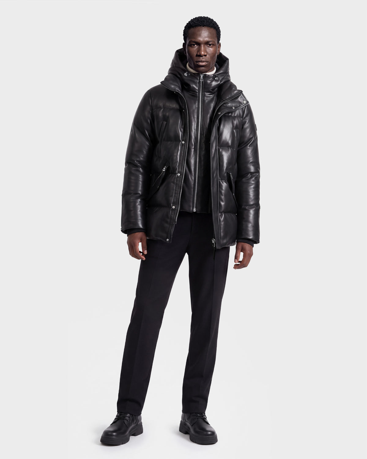 A man stands against a plain background wearing the Rudsak APOLLO X - 6125696 Black leather puffer jacket, paired with black pants, a black shirt, and black shoes.